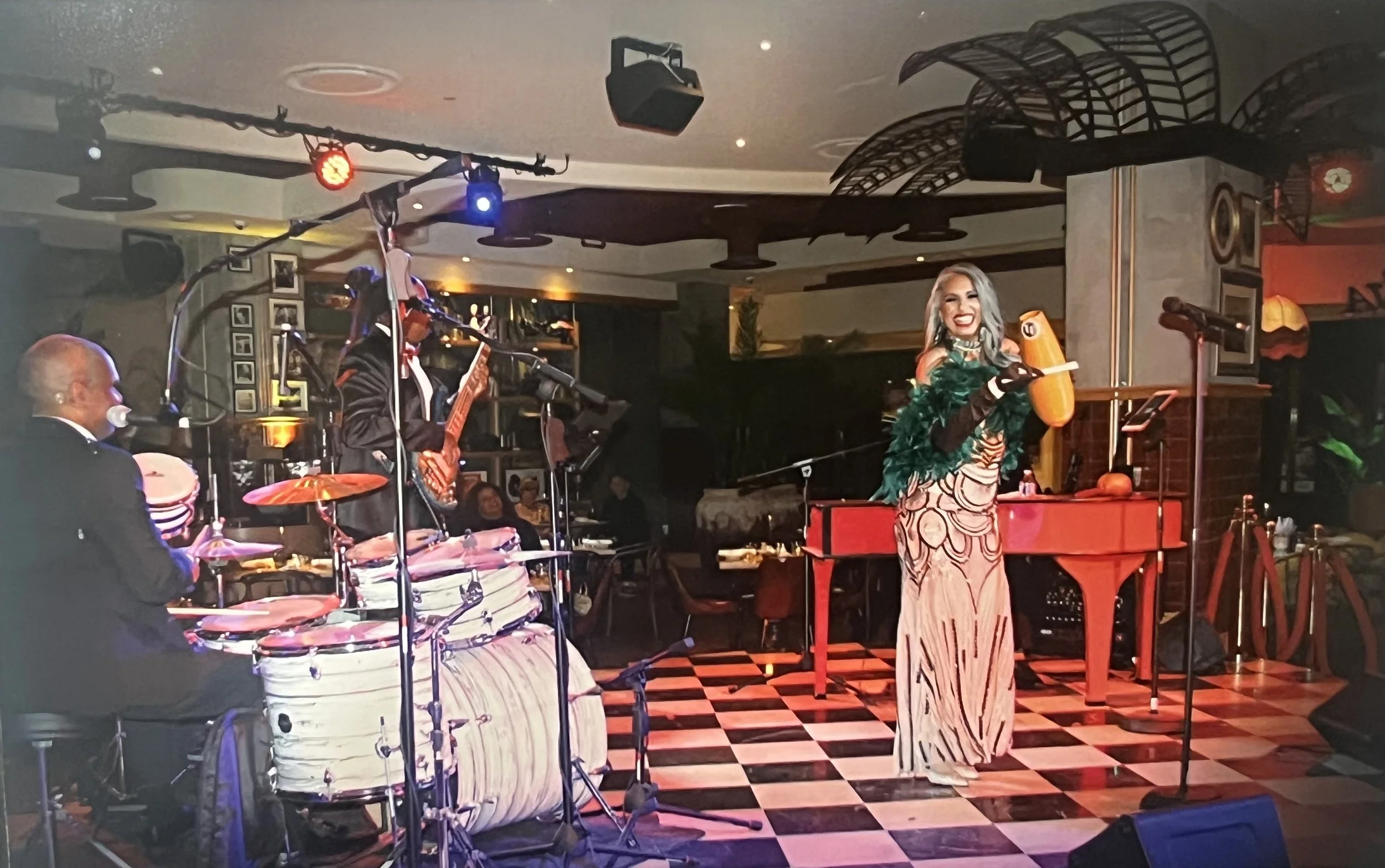 Live jazz band performing in a dimly lit venue, with a woman singer holding a microphone and a percussionist playing drums. The setting has a checkered floor and warm ambient lighting.