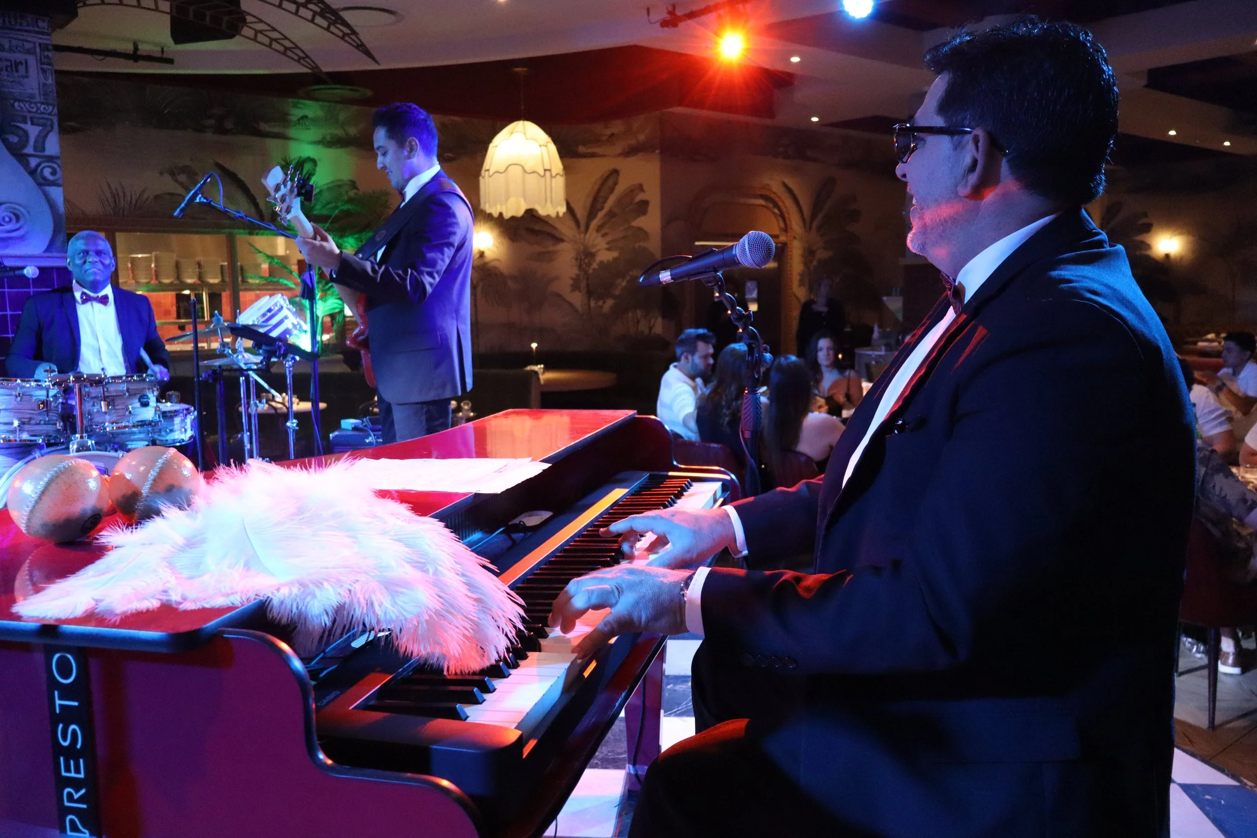 Musicians performing live on an indoor stage, with a pianist and a guitar player in the foreground, and a drummer in the background, with audience seated at tables in a dimly lit venue decorated with tropical painting murals.
