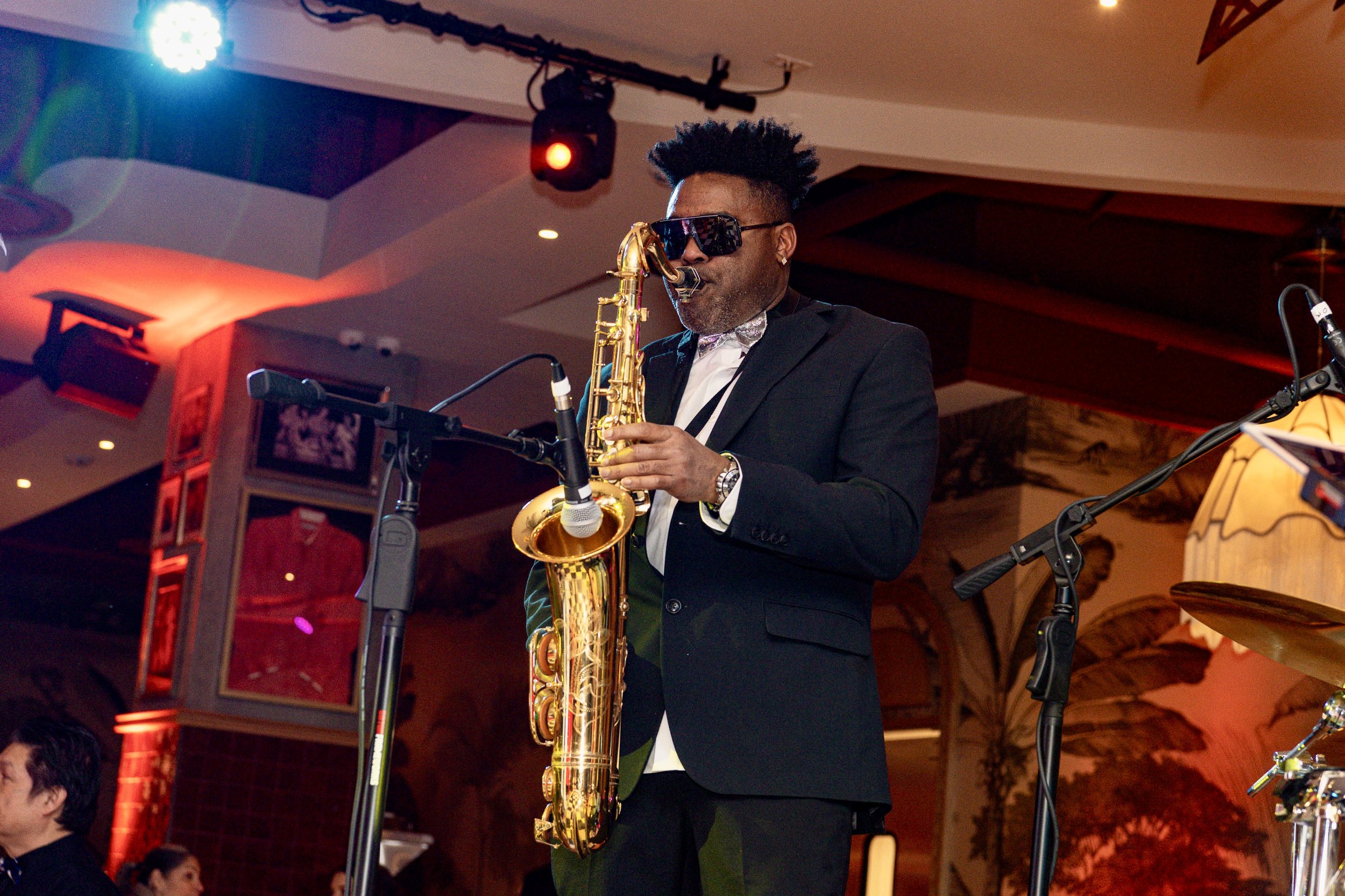 A man wearing a black suit, white shirt, and dark sunglasses playing a gold saxophone on stage during an indoor event.