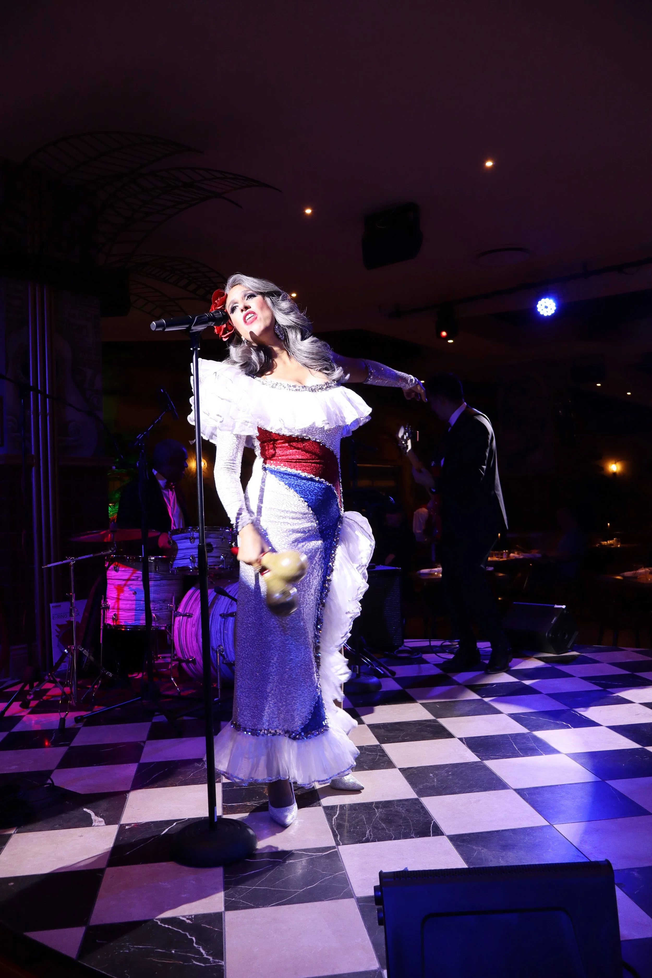 A woman dressed in a vintage-style dress performs on stage with a vintage microphone in her hand, surrounded by musicians. The scene is lit with colorful stage lighting, and the stage has black and white checkered flooring.