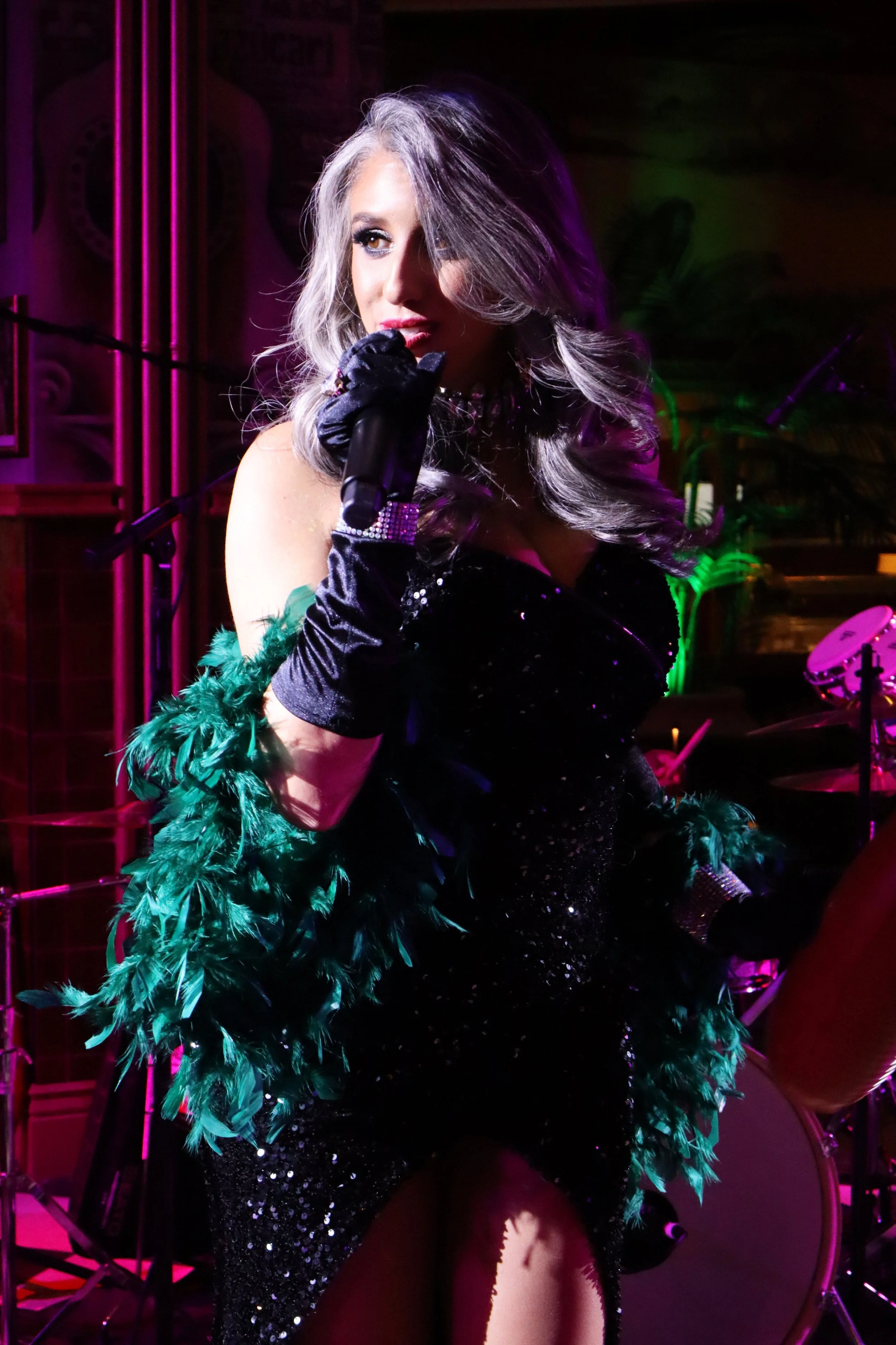 A woman dressed in a black, sparkling dress with green feathered arm accessories, holding a microphone, performing on stage with colorful lighting.