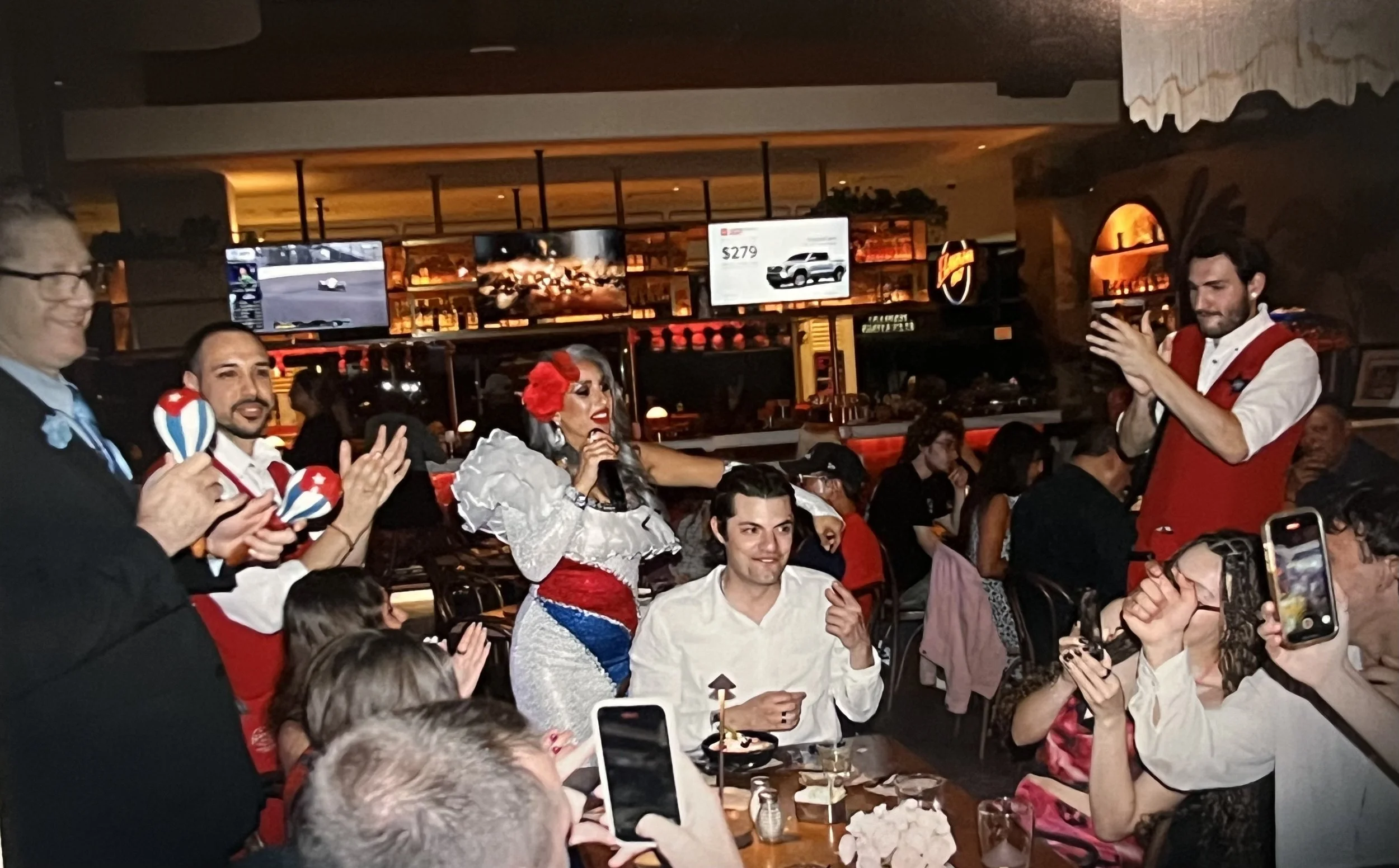 People celebrating at a restaurant or bar, featuring a performer dressed in a white costume with red, white, and blue accents, holding a microphone and singing, while others around are taking photos and clapping.