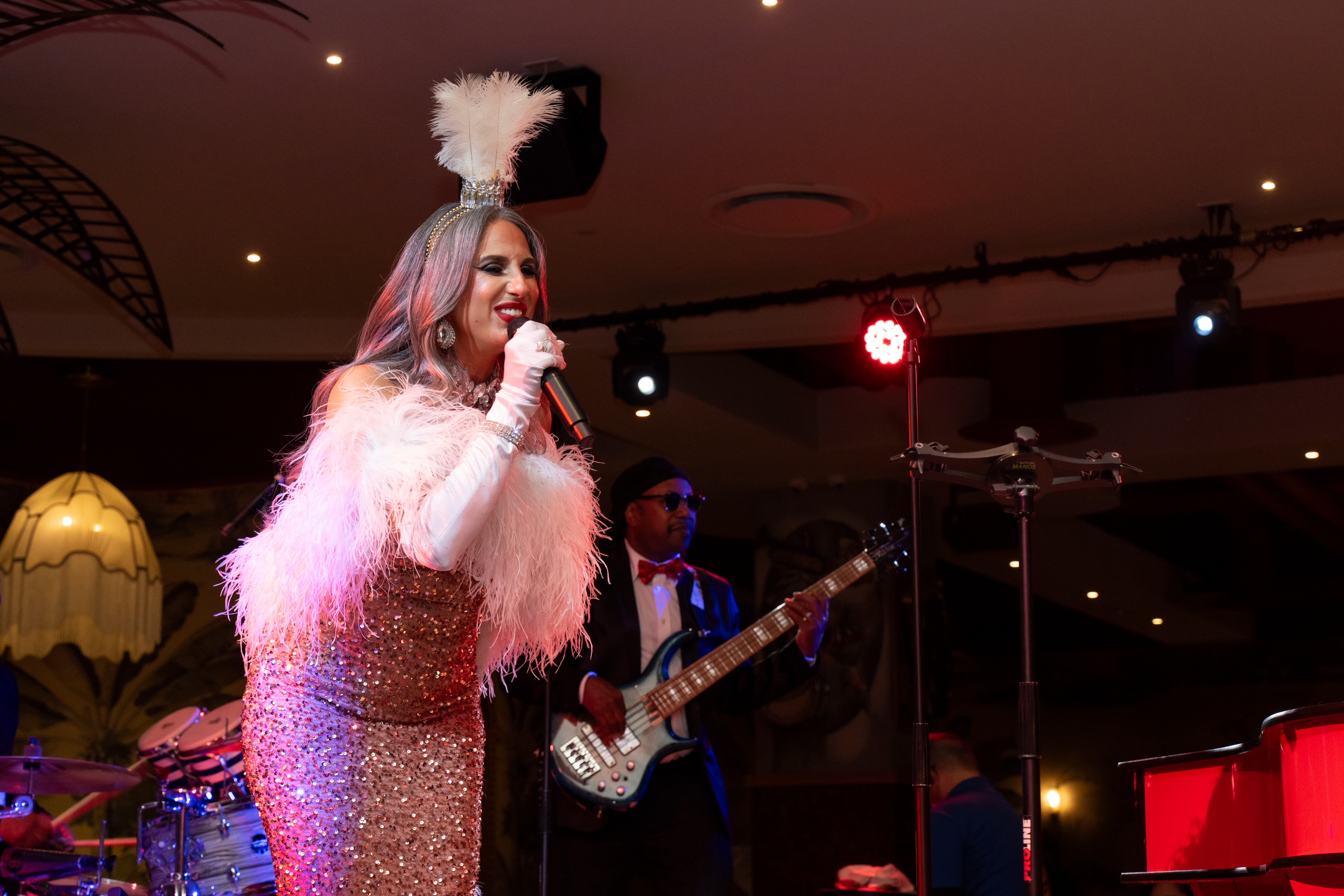 A female singer dressed in a glittery pink gown with feathered embellishments and a feather headpiece, singing into a microphone, accompanied by a male bassist wearing sunglasses and a suit at an indoor performance venue with stage lighting.