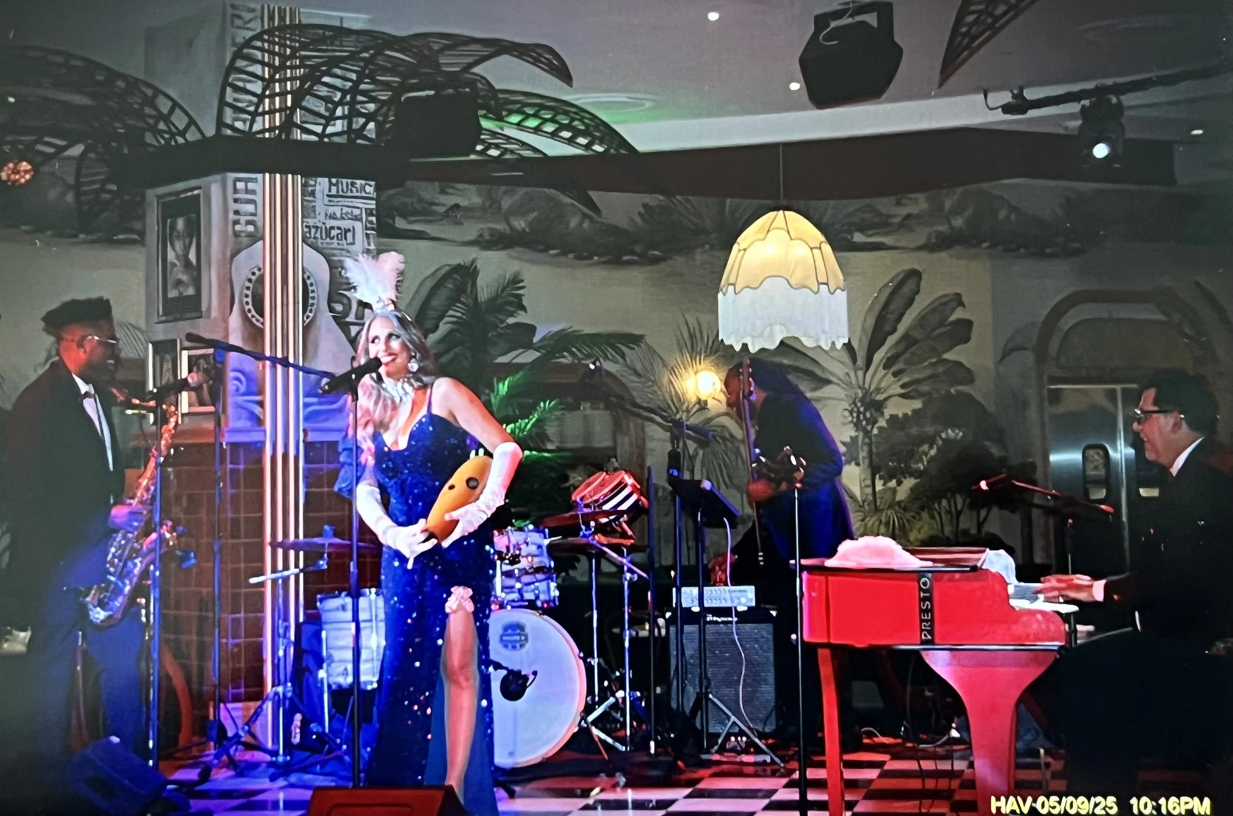 Musicians performing on a small stage: a woman in a sparkly blue gown with a feathered headpiece holding a guitar, a man on the left playing saxophone, a woman in the background playing a percussion instrument, and a man on the right playing a keyboa