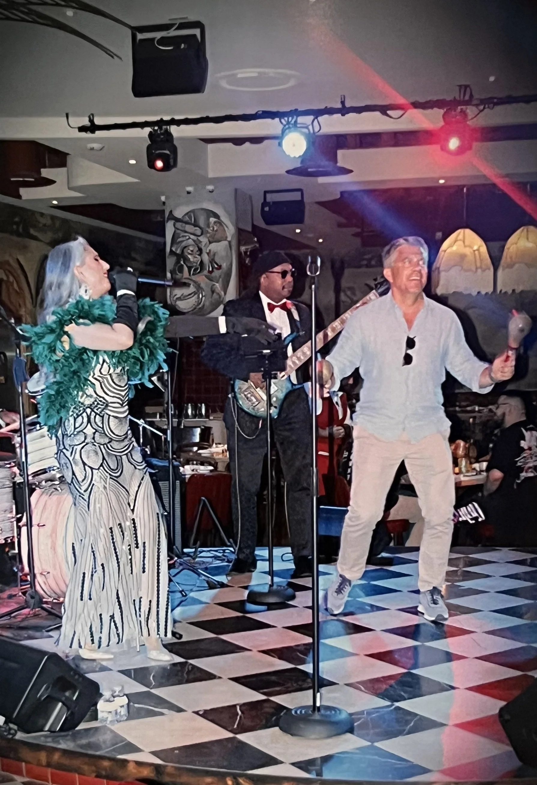 A band performing on stage with a woman singing into a microphone, a man playing the guitar, and another man dancing and singing with maracas, in a lively indoor setting.