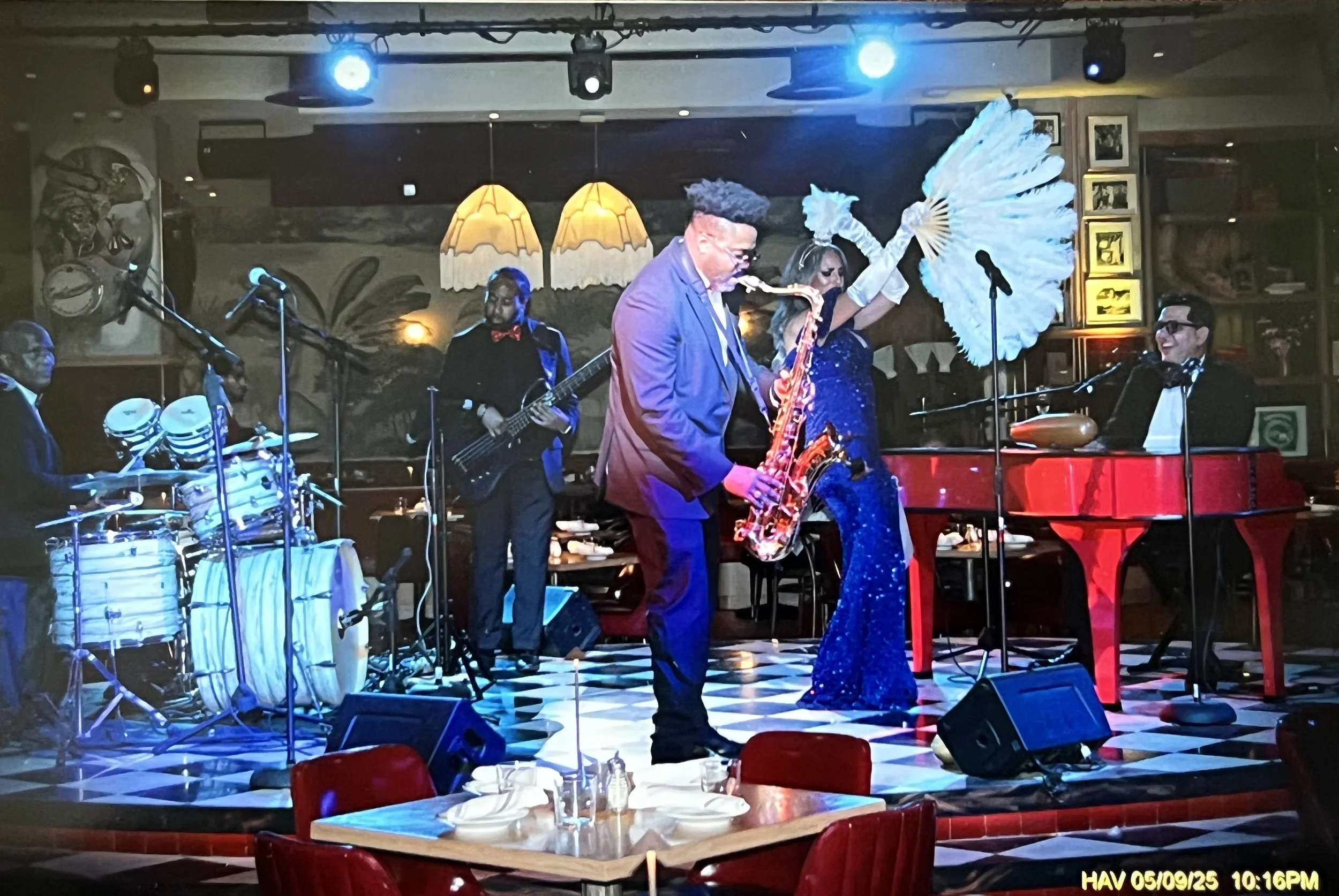 live jazz band performing on a stage at a restaurant. Musicians include a drummer, a bassist, a saxophonist, a pianist, and a singer with large white fan and holding a fan. The scene features colorful lighting, black-and-white checkered floor, and ta