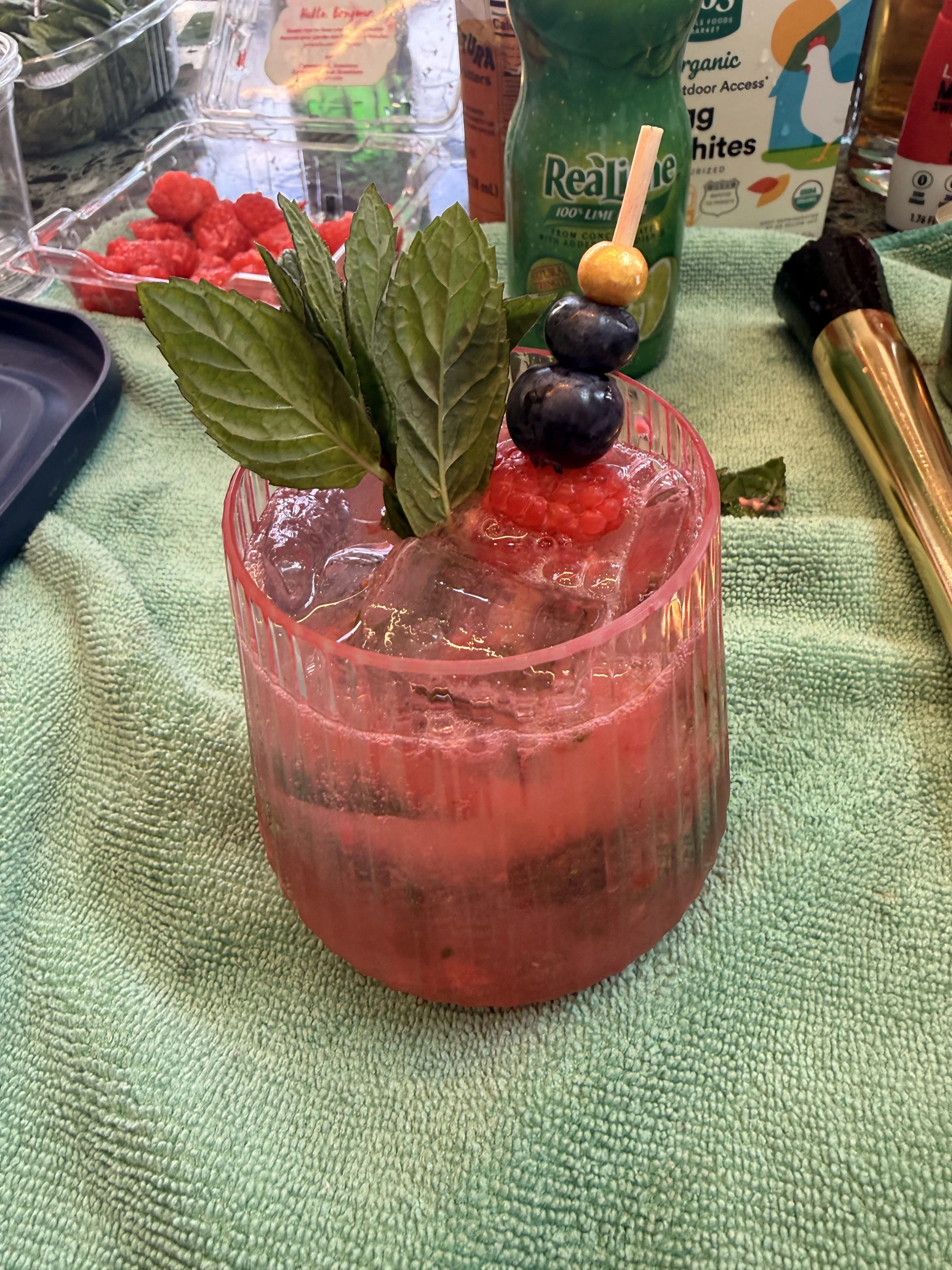 A pink cocktail garnished with fresh mint leaves, a skewer with blueberries and a melon ball, served over ice in a pink glass, on a green textured towel surrounded by various drink ingredients and tools.