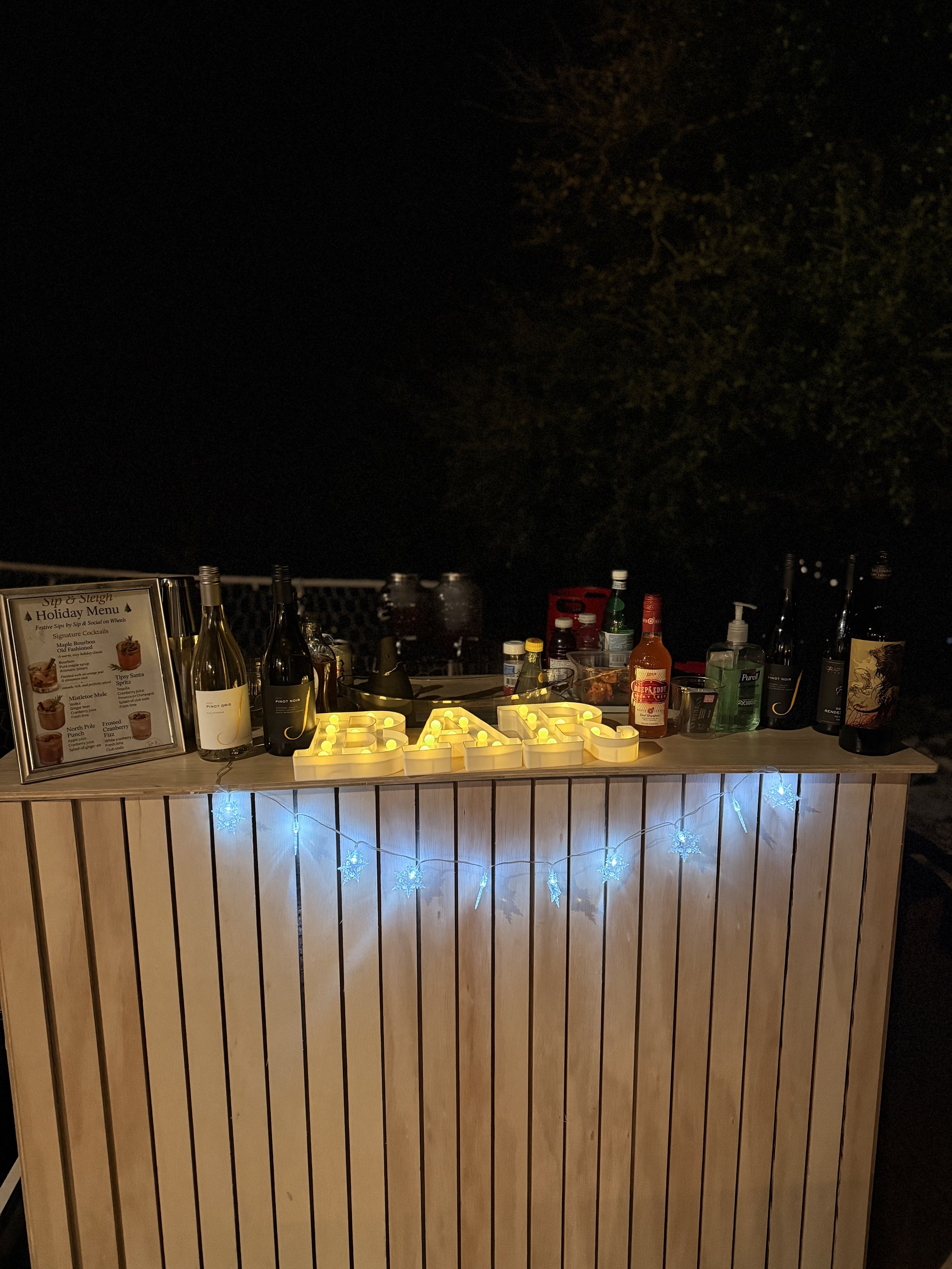 A bar set up outdoors at night with illuminated yellow letters spelling BAPS, string lights with snowflake shapes, and bottles of alcohol and mixers on the counter.