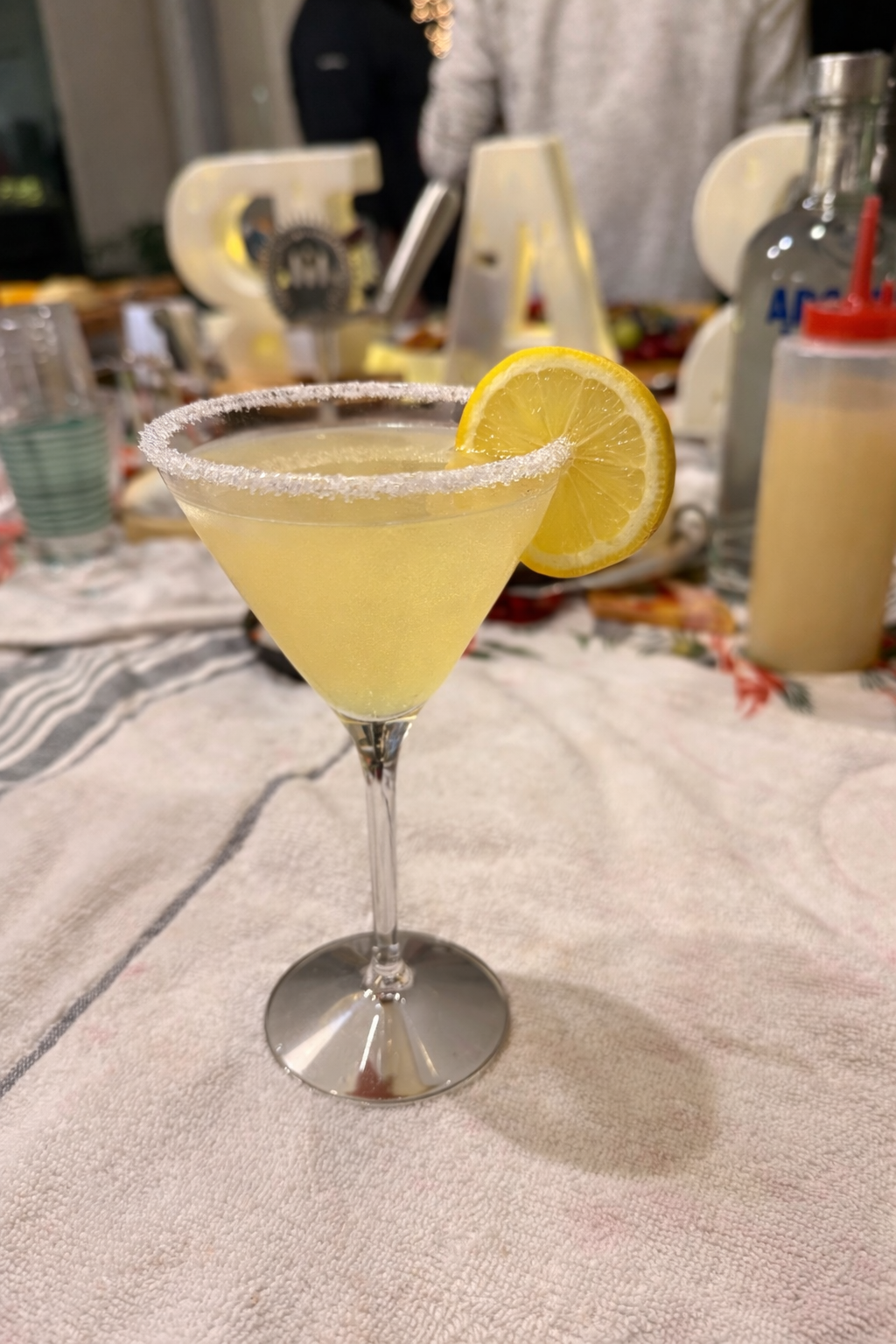 A yellow cocktail in a martini glass with a sugared rim, garnished with a lemon slice, on a table with a floral tablecloth. In the background, out of focus, there are decorative letters and cocktail supplies.