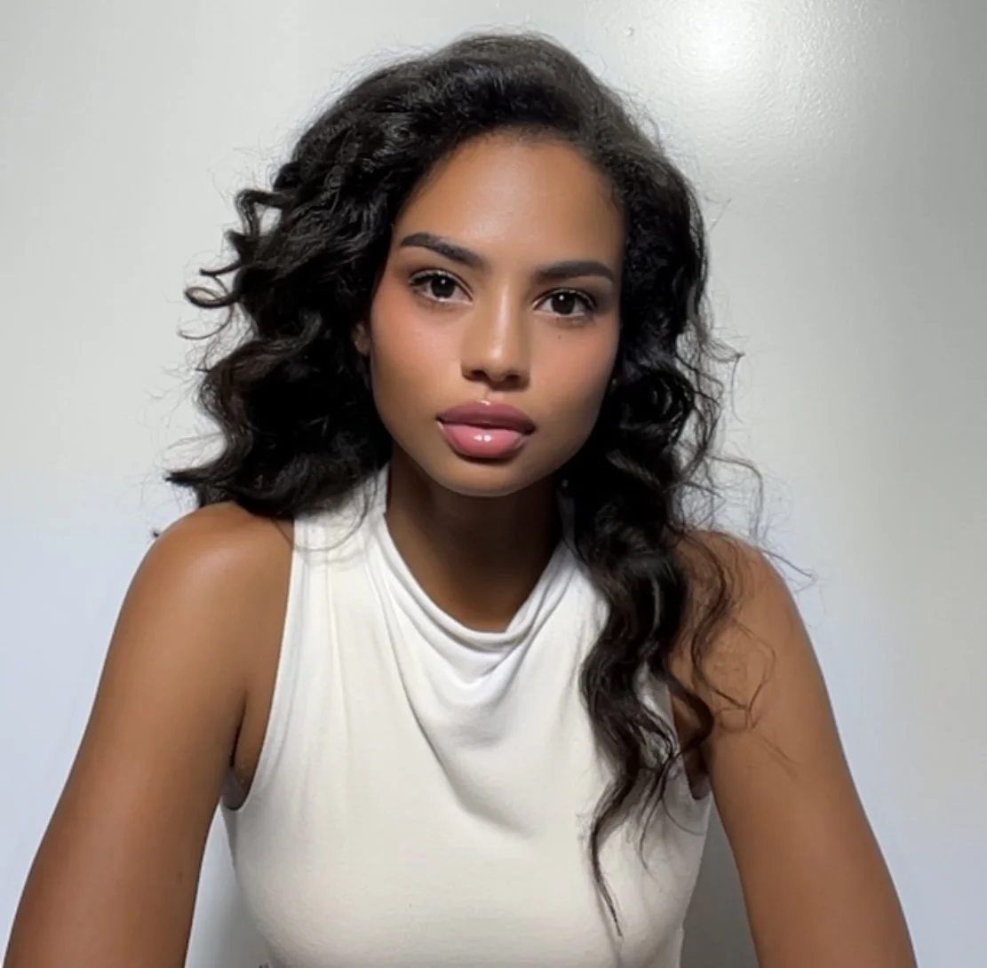 A young woman with curly black hair, wearing a sleeveless white top, looking directly at the camera with a neutral expression, in front of a plain light gray background.
