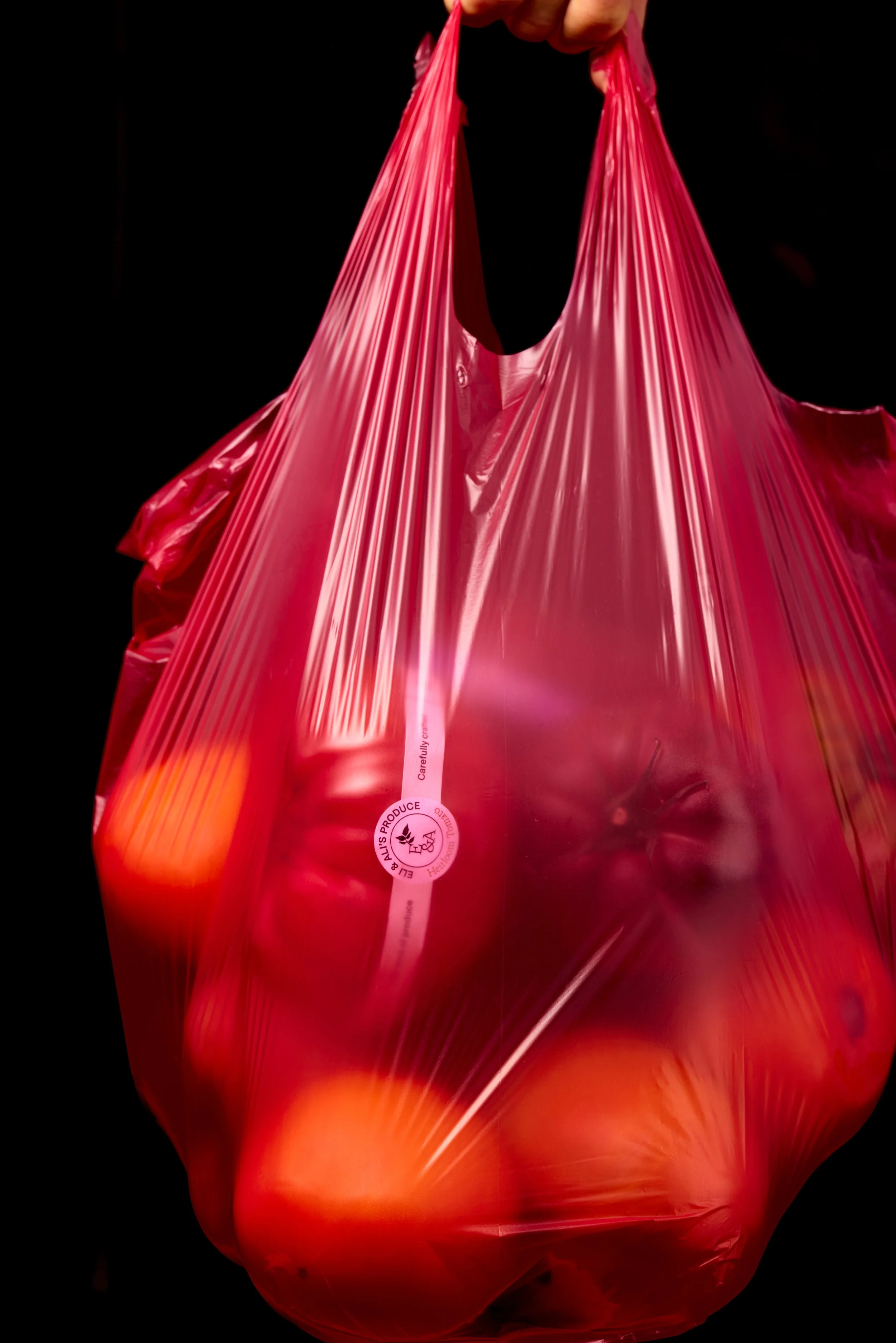 Red plastic shopping bag containing tomatoes. The bag is held by a hand against a black background.