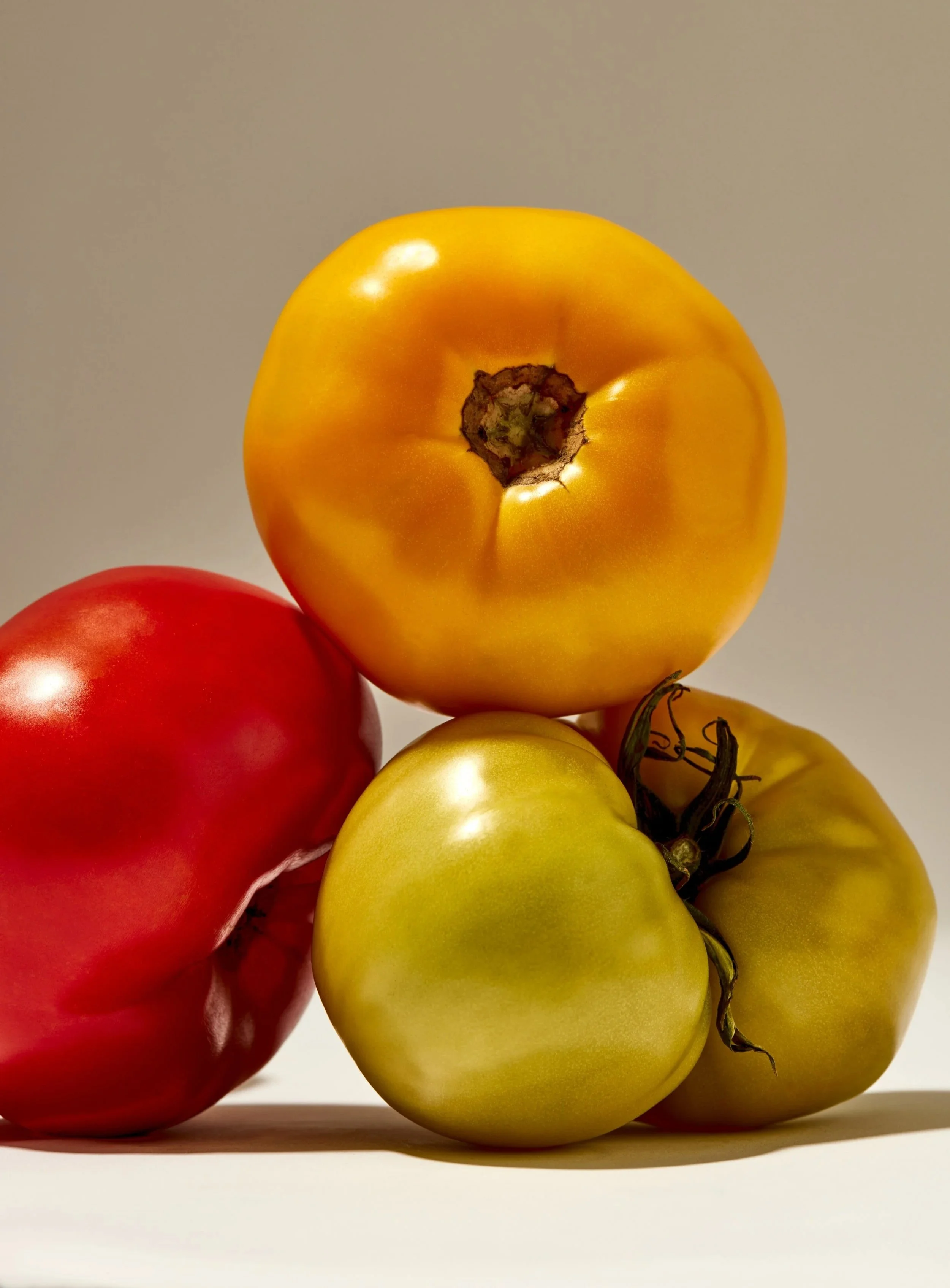 Close-up of four hanging tomatoes, yellow and red, with the yellow one on top and the red one at the bottom left, against a plain gray background.
