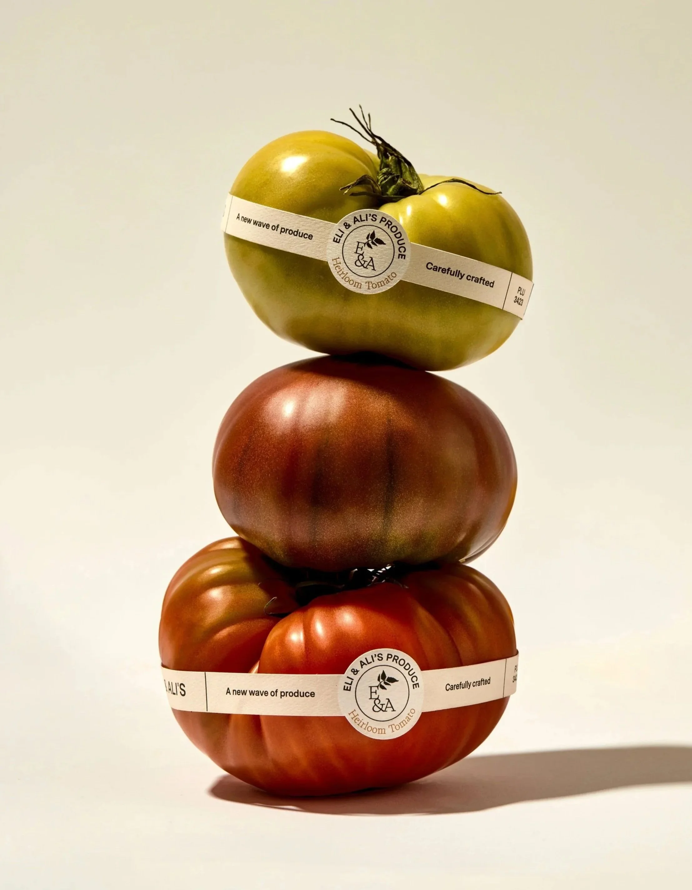 Stacked heirloom tomatoes of different colors with produce labels wrapped around each, set on a plain white background.