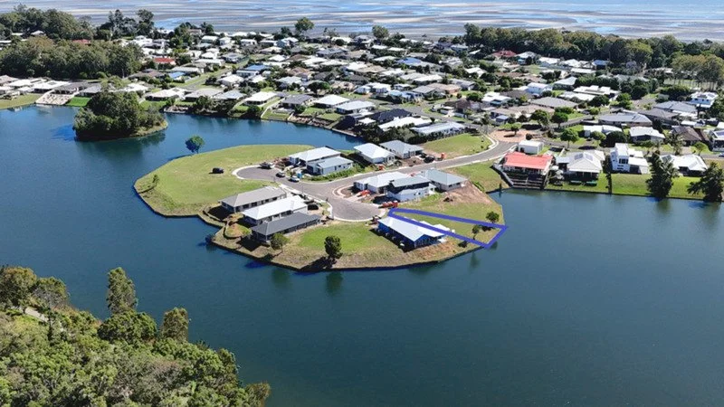 Aerial view of a residential neighborhood on a small island with houses, a body of water surrounding it, and a marina or boat dock area outlined in blue.