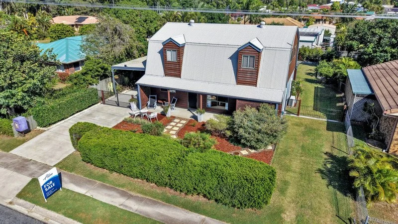 A two-story house with a metal roof, a front porch with seating, surrounded by a well-maintained lawn and shrubbery, with a 'For Sale' sign in the yard.