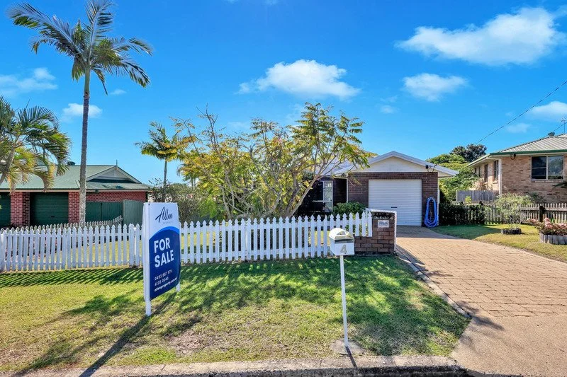 Single-story house with a white picket fence, a for sale sign, a mailbox, and a driveway on a sunny day with blue sky and scattered clouds.