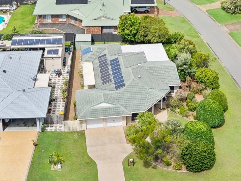 Aerial view of a house with a green roof and solar panels, surrounded by green trees and neighboring houses with solar panels on their roofs.