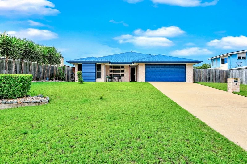 Front view of a blue house with a garage, a driveway, a front lawn, a wooden fence, and neighboring houses under a partly cloudy sky.