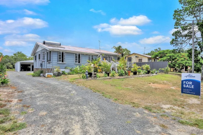 A house with light blue siding, surrounded by a gravel driveway and a lawn with plants, for sale sign, and a cloudy blue sky.