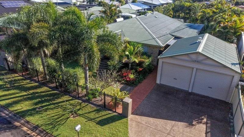 Aerial view of a backyard with lush green grass, palm trees, and a garden with flowering bushes, fenced with a black metal fence, next to a two-car garage and neighboring rooftops.