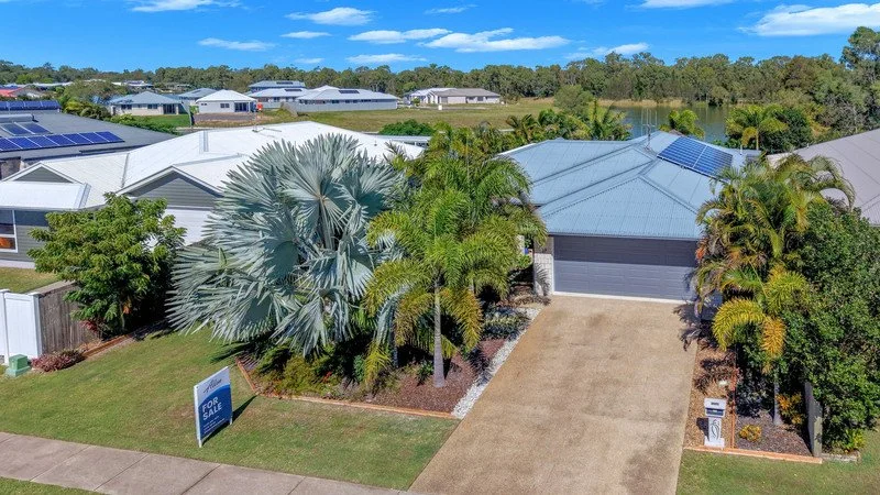 A single-story house with solar panels on the roof, surrounded by lush green trees and shrubs, with a driveway leading to a garage, and a 'For Sale' sign on the lawn.