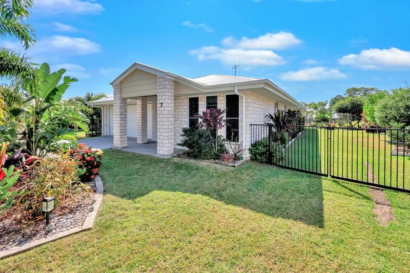 A single-story house with a white brick exterior, surrounded by a lush green lawn and garden beds, under a blue sky with some clouds.