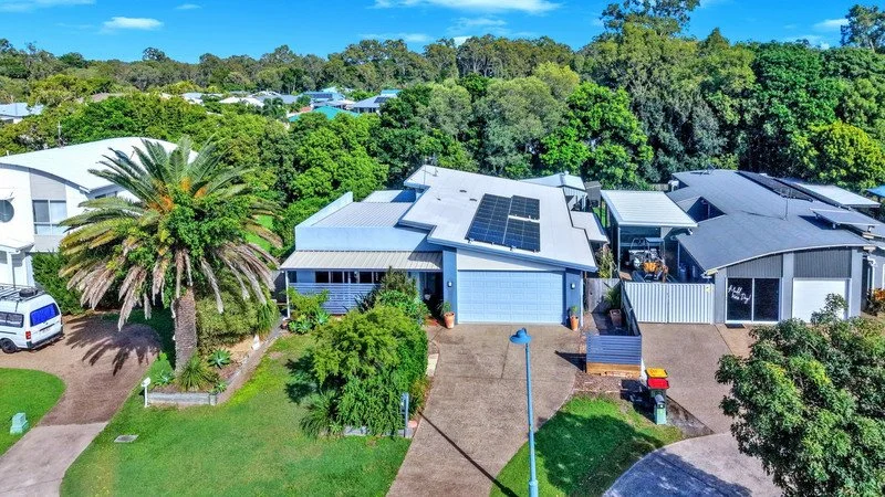 Aerial view of modern houses with solar panels on roofs, surrounded by green trees and lawns, with a clear blue sky overhead.