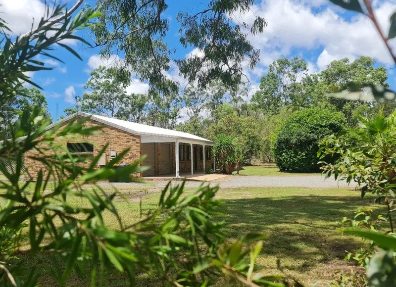 A house with a white roof is surrounded by green trees and bushes under a blue sky with clouds.