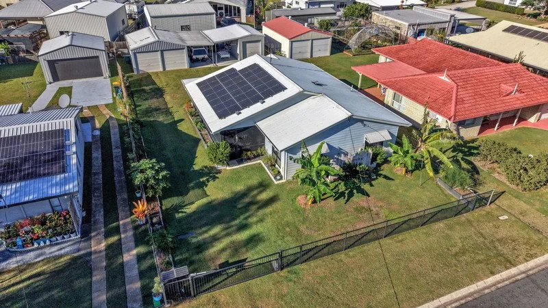 Aerial view of a residential backyard with solar panels on the roof, a greenhouse with solar panels on top, and neighboring houses with solar panels, landscaped lawns, and palm trees.