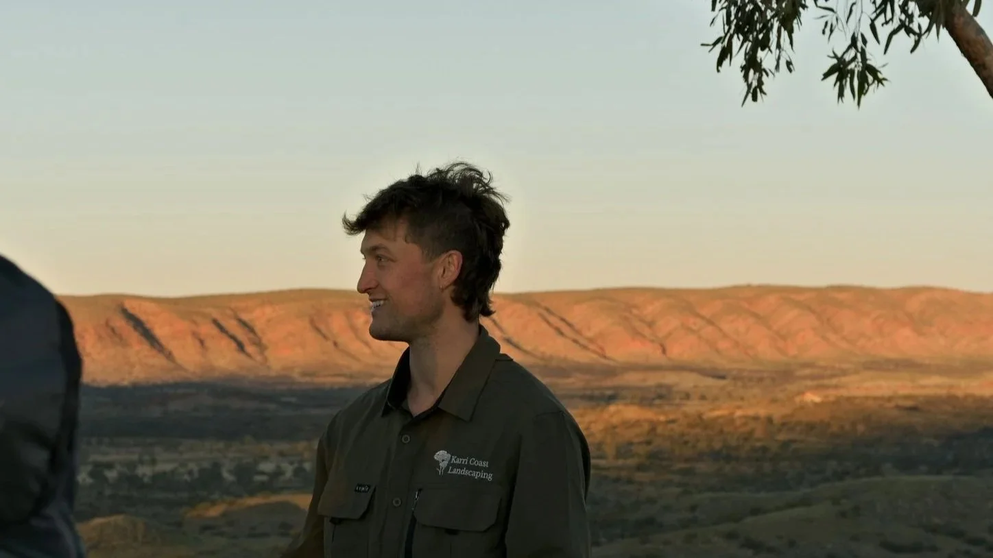 A man standing outdoors with a landscape of mountains and open land at sunset in the background, wearing a dark green shirt with a logo for Karr-Coast Landscaping.