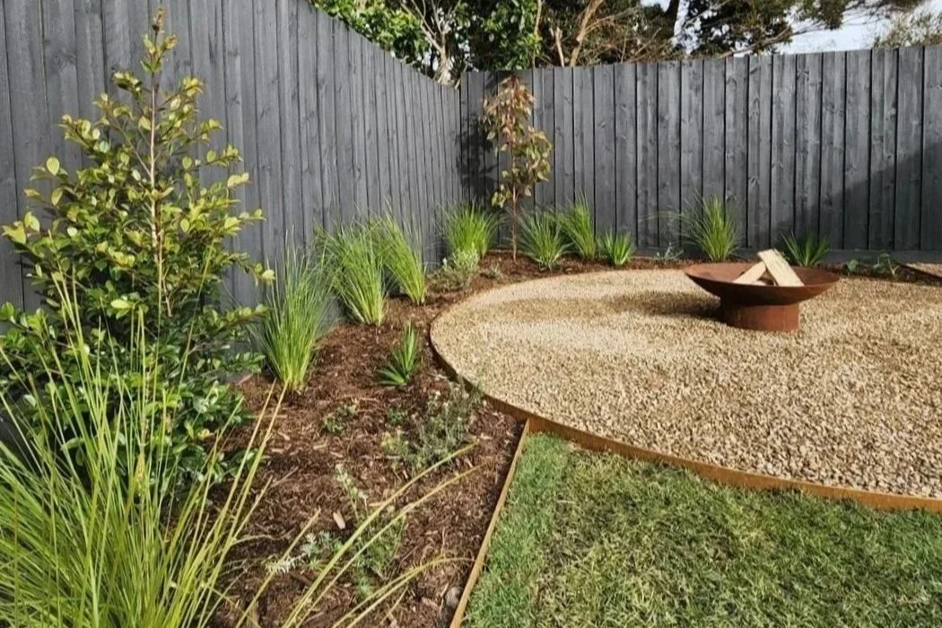 Backyard garden with green plants, steel edging, small trees, a black wooden fence, and a firepit in a gravel area and some grass.