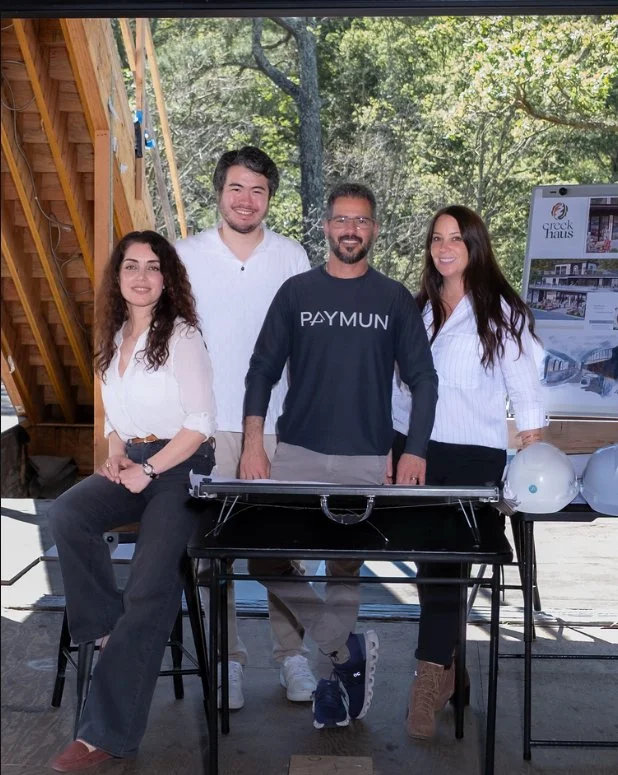 Four people standing behind a black table with a blueprint on it, outdoors with trees in the background. Two men and two women, casually dressed, smiling at the camera. One woman is seated next to the table, and there are construction helmets on the table.