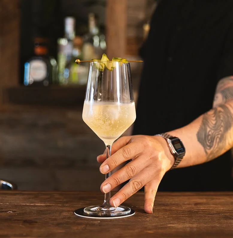 A person with a tattooed arm wearing a black shirt and a smartwatch is holding a wine glass filled with a fizzy, pale yellow cocktail garnished with a skewer of twisted lemon peel, on a wooden bar counter.