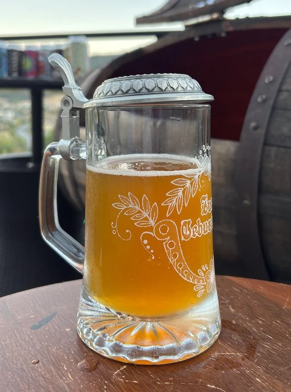 A glass beer mug with a decorative design, filled with light amber beer, sitting on a wooden table outdoors with a large barrel in the background.