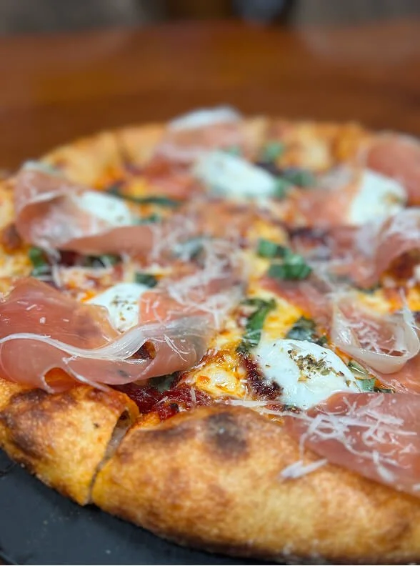 Close-up of a prosciutto and basil pizza with melted cheese and herbs.