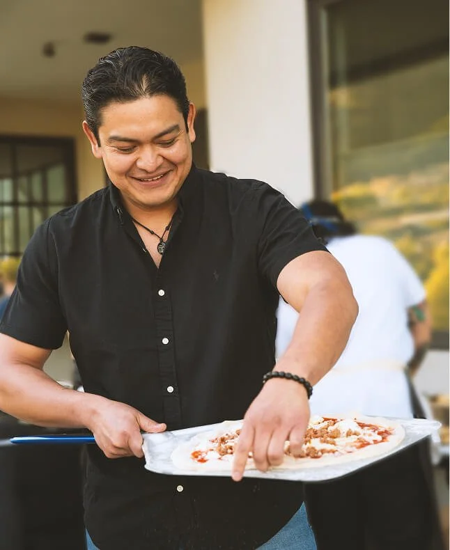 A smiling man with dark hair, wearing a black shirt and a beaded bracelet, holding a pizza on a tray outdoors.