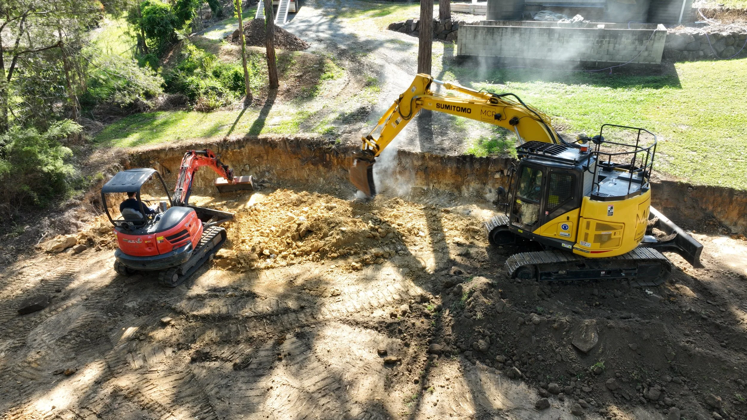 Construction site with two excavators doing site preparation. A large yellow excavator is scooping soil, while a smaller red excavator is nearby. The area is surrounded by trees and greenery.