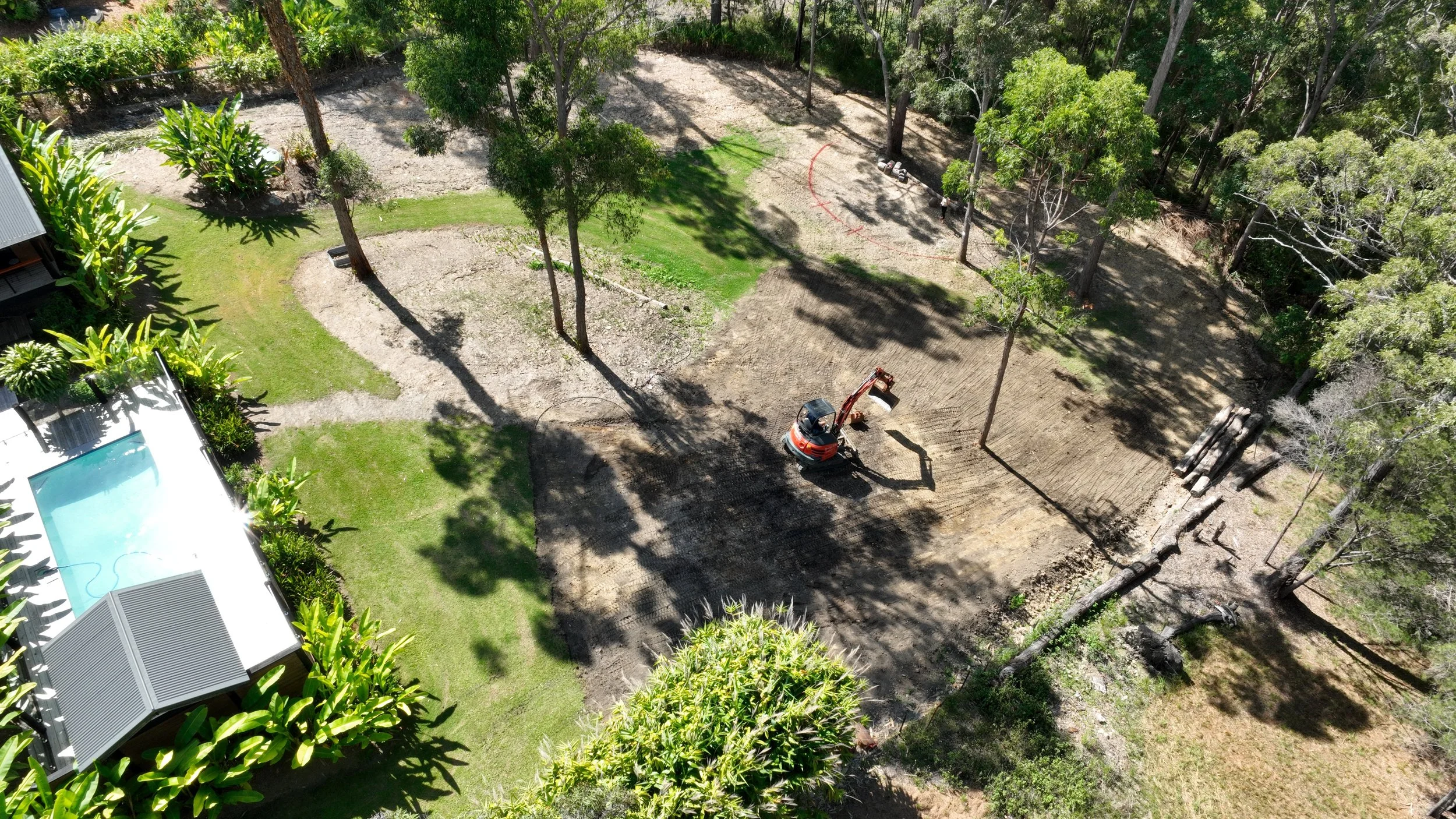 Aerial view of a backyard with a swimming pool, green grass, trees, showing an excavator doing site clearing. 
