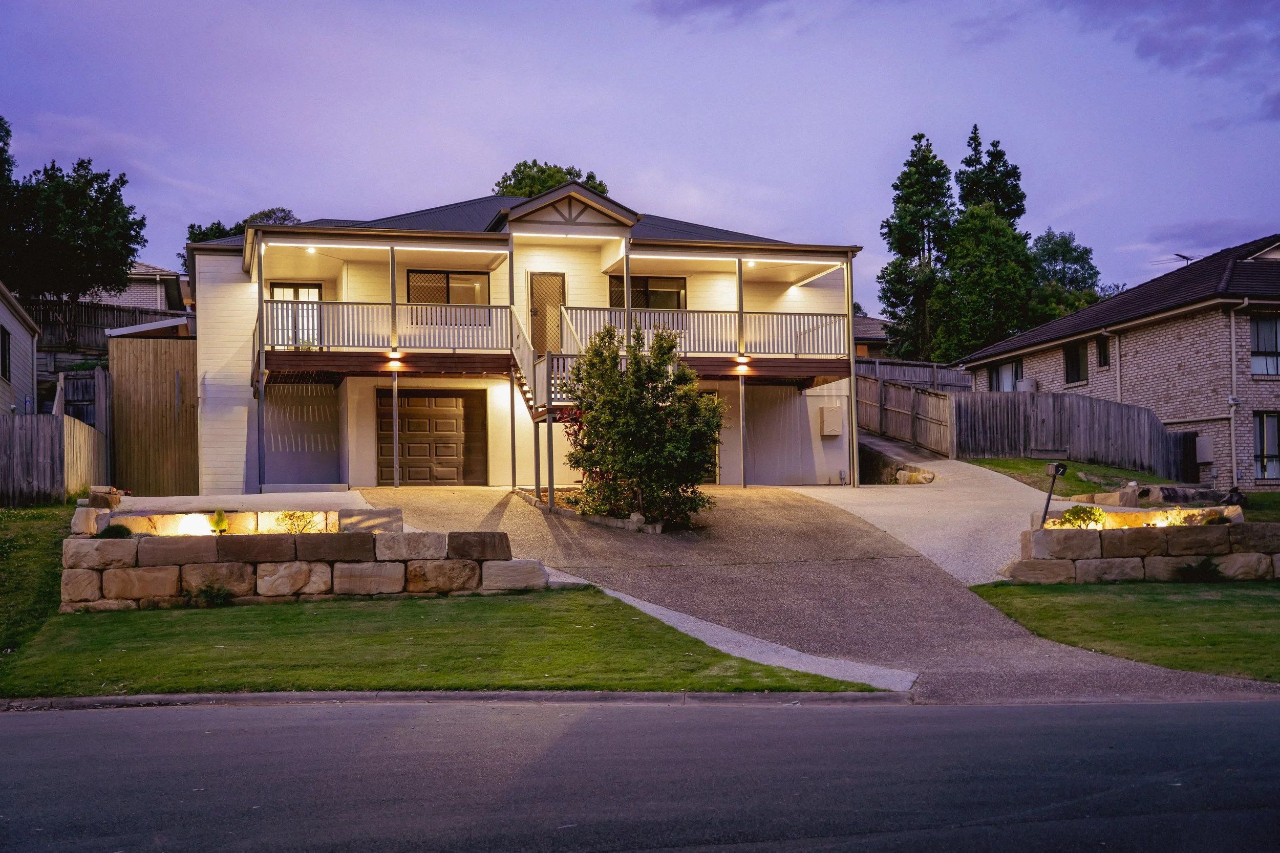 A two-story house illuminated at dusk, with a front yard, stone retaining wall, and driveway, surrounded by neighboring houses and trees.
