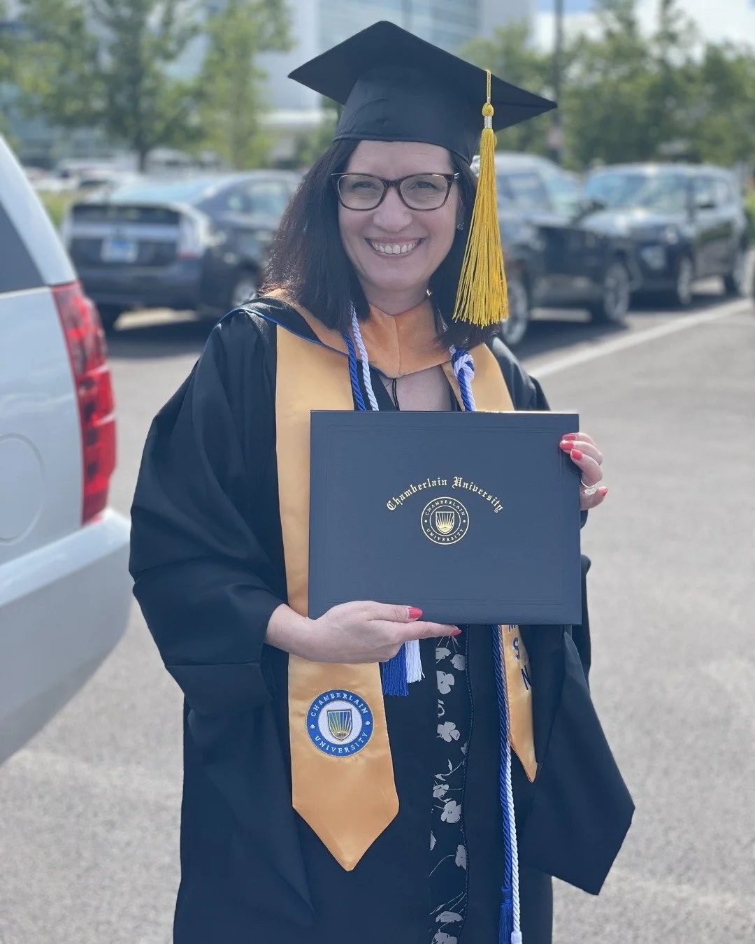 Danielle Penman in graduation gown and cap smiling while holding a diploma from Chamberlain University, outdoors in a parking lot.