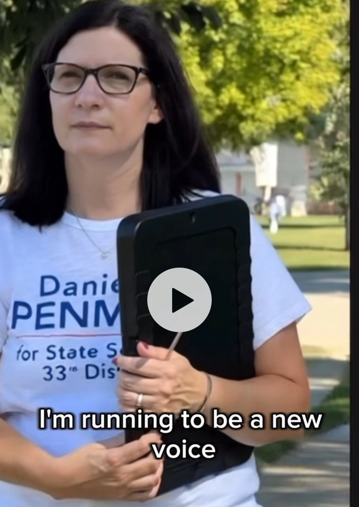 Danielle Penman wearing a white campaign T-shirt holding a clip board and other people in the background, with subtitles reading 'I'm running to be a new voice'.