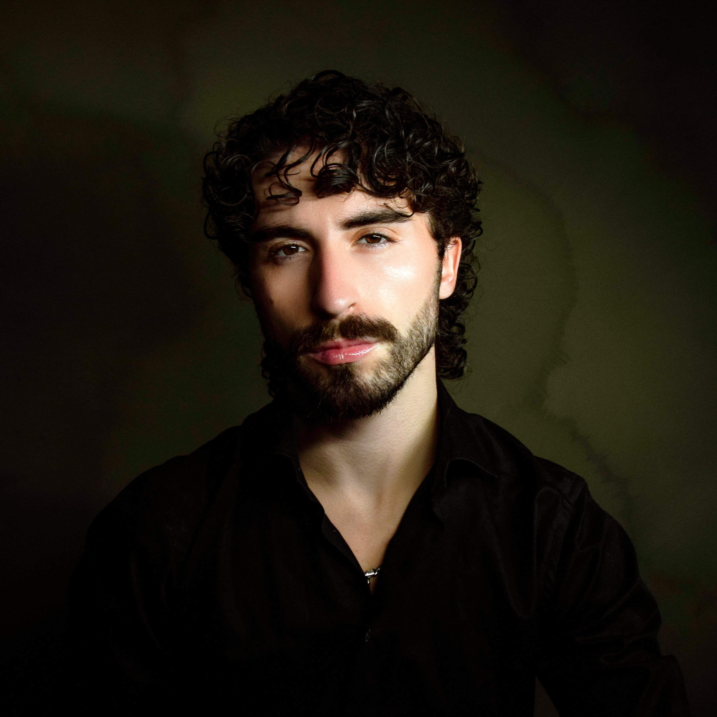 Close-up of Stephen Marrello. Curly dark hair, beard, and mustache, wearing a black shirt against a dark background.