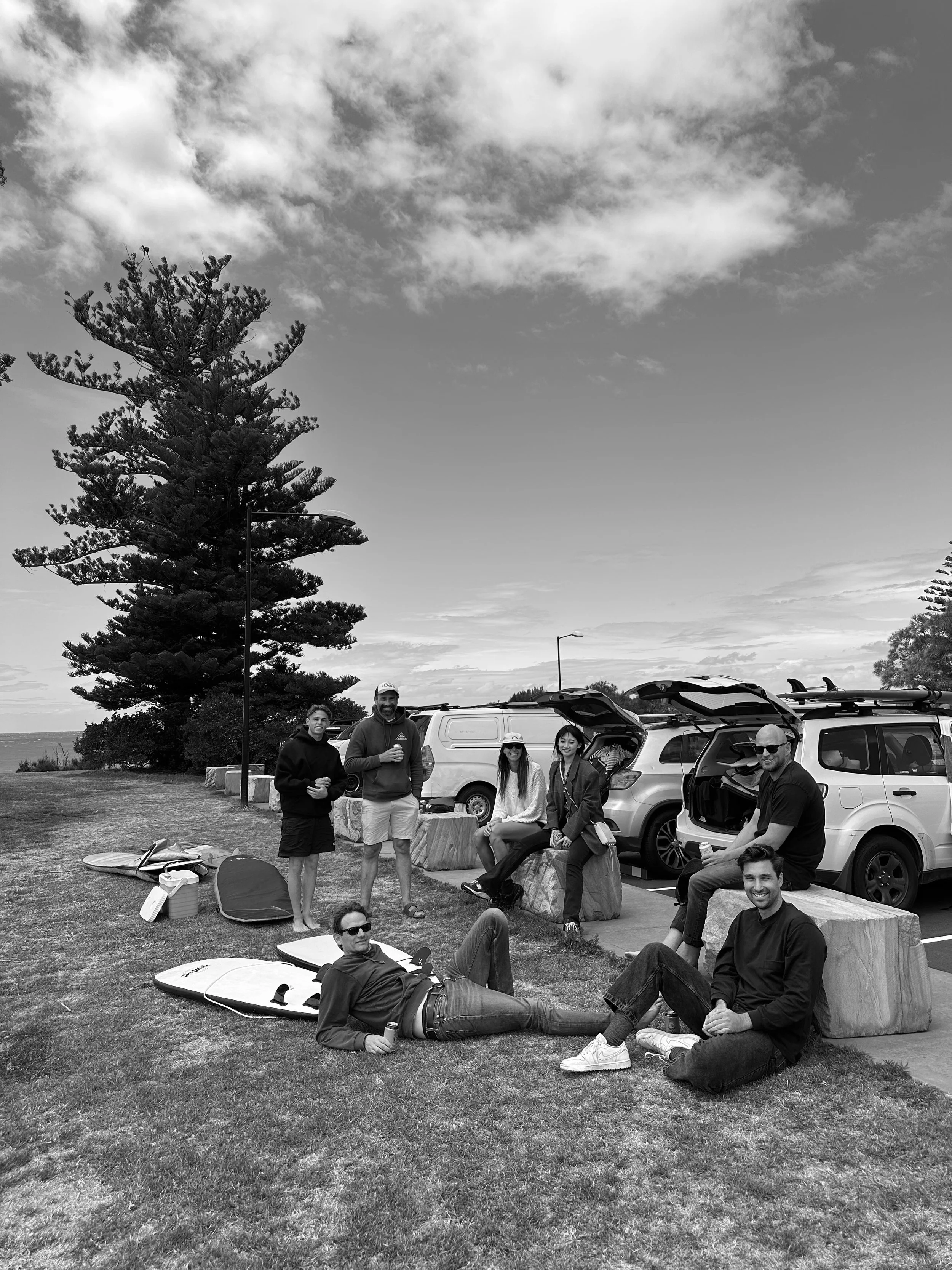 A group of seven people hanging out at a park with cars parked behind them and surfboards on the grass.