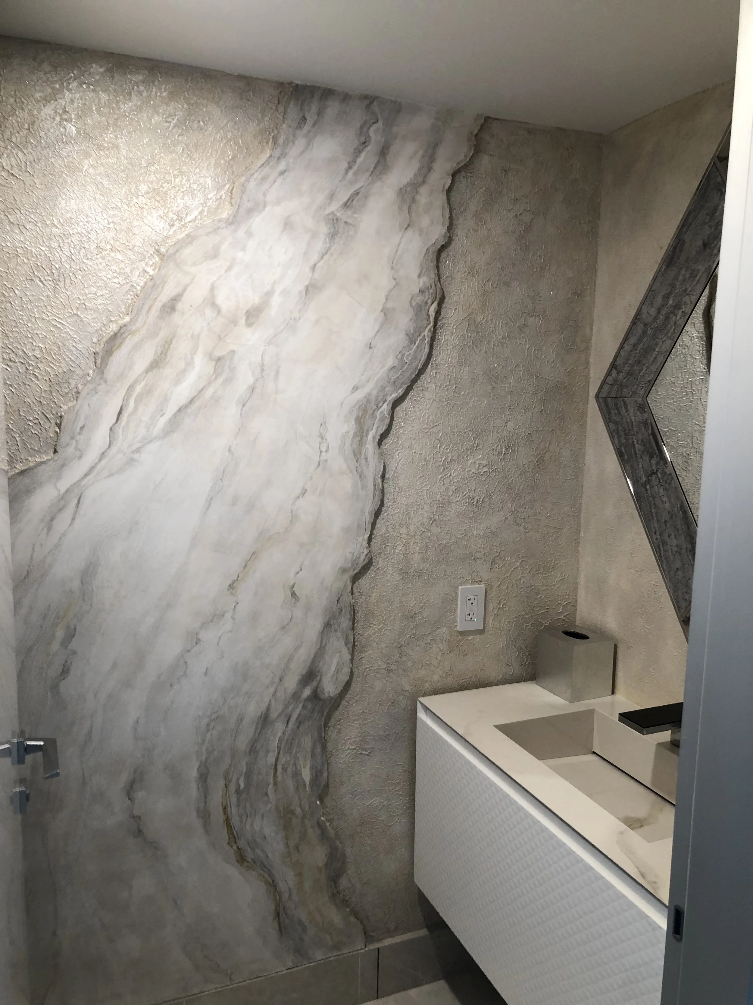 A modern bathroom corner with textured beige walls, a large marble-style accent wall, a wall-mounted white sink, and a dark framed mirror.