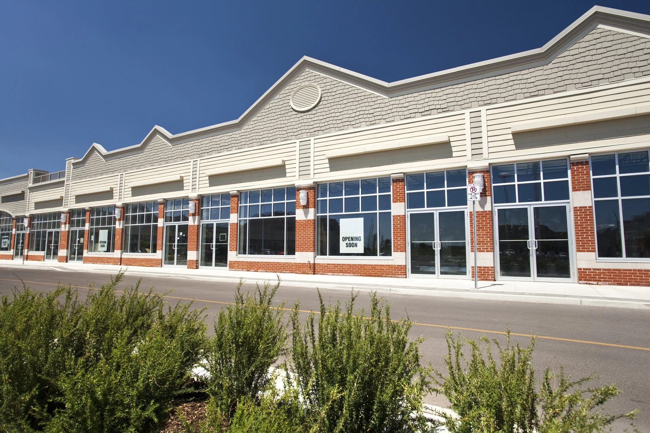 Empty retail plaza with large glass storefronts, some with signs reading "opening soon," under a clear blue sky.