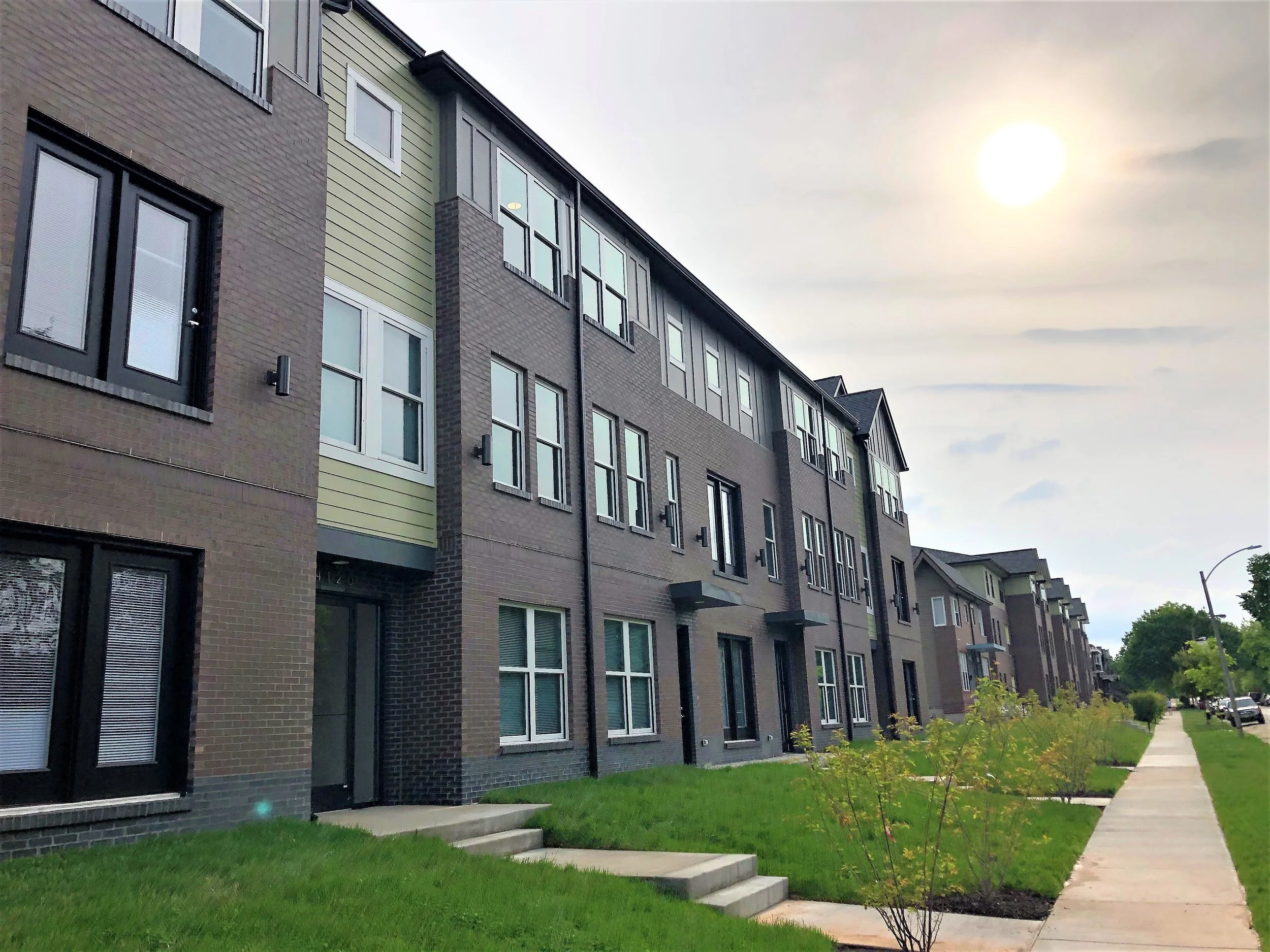 A row of modern multi-story townhouses with brick and siding exteriors, set along a sidewalk with small bushes and grass, under a cloudy sky with the sun visible.