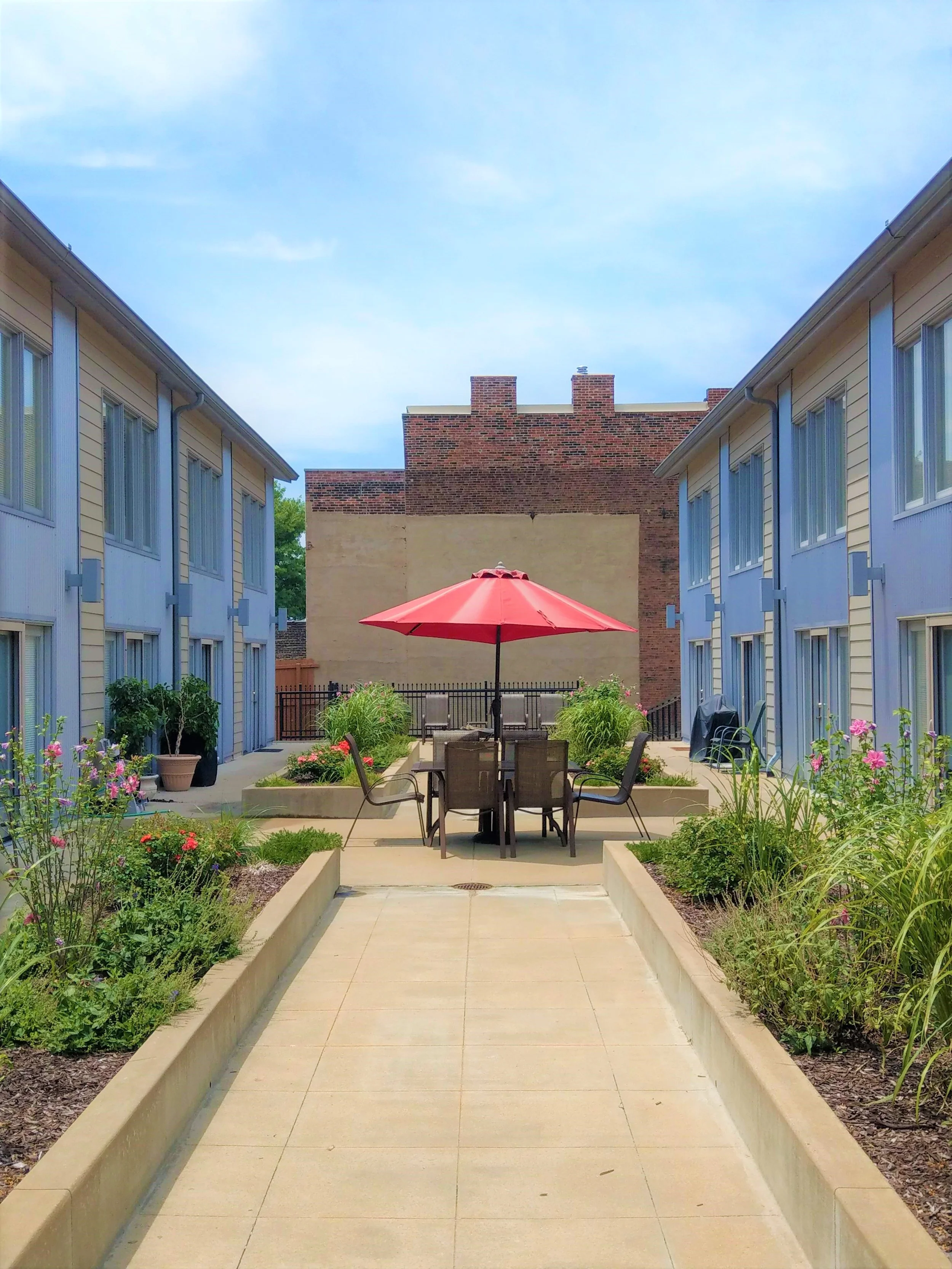 An outdoor courtyard with a round table and six chairs around a red umbrella, surrounded by garden beds with plants and flowers, flanked by two yellow buildings with blue trim.