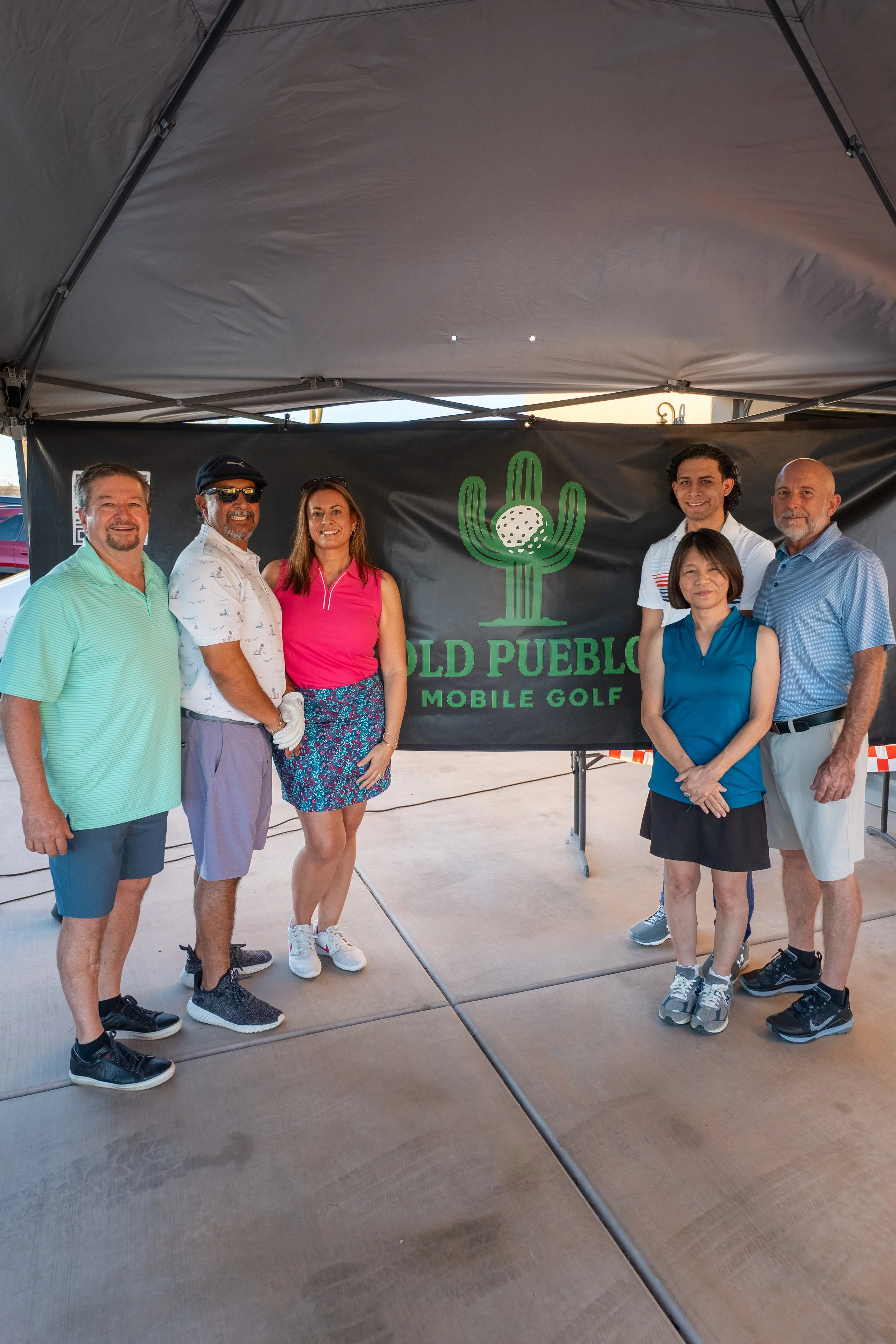 Group of seven people standing under a canopy with a banner that reads 'Old Pueblo Mobile Golf' and features a cactus logo.