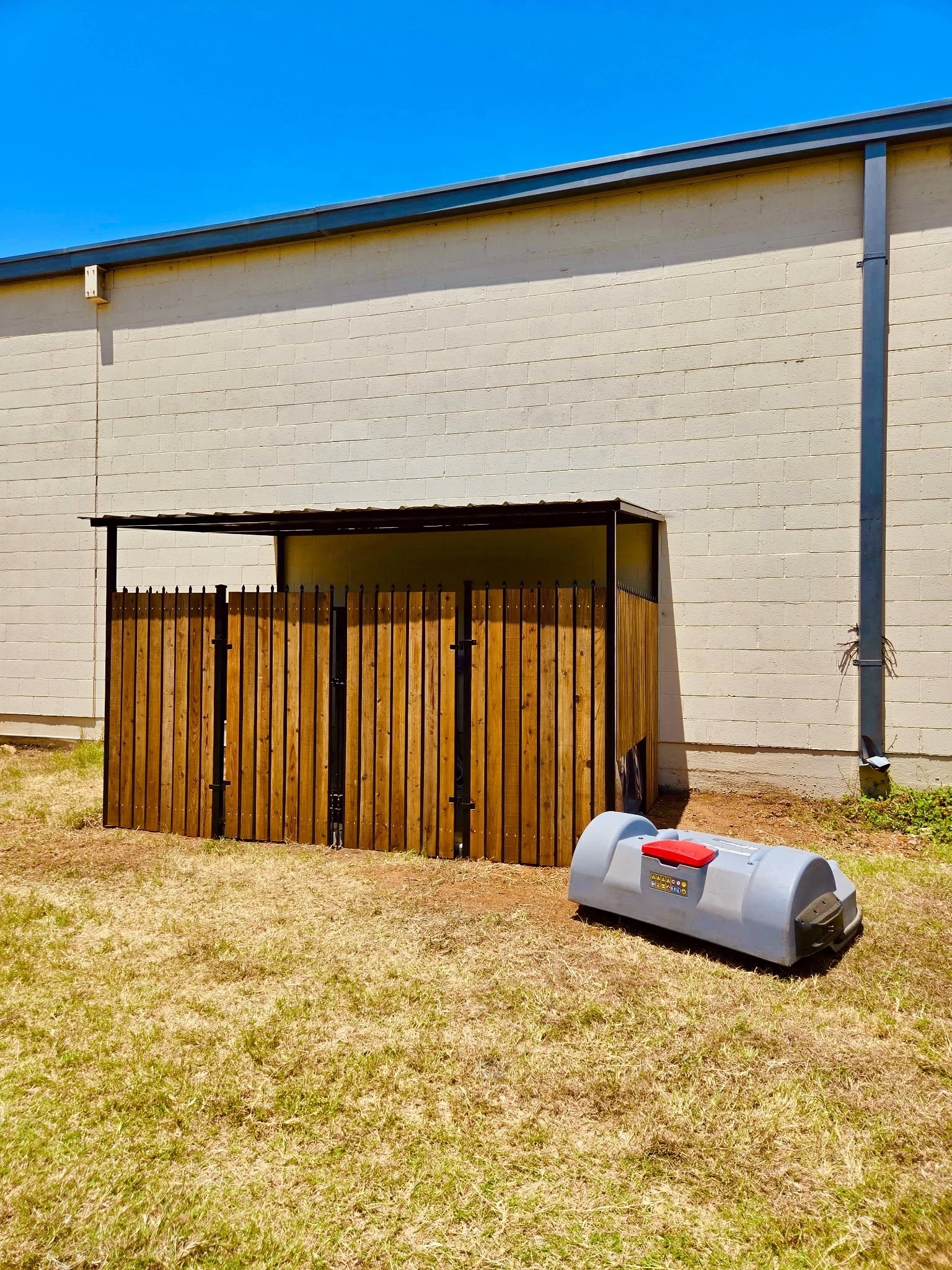 Backyard with a white brick wall, a wooden fenced structure, a grey robotic lawn mower, and a blue sky.