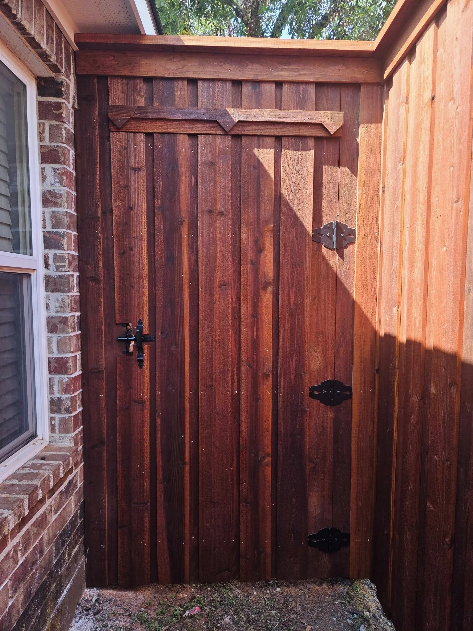 A newly installed wooden fence with a decorative top, secured with black metal hinges and a latch, standing next to a brick house with a window on the left. The ground in front of the fence has dirt and small plants, and a tree with green leaves is v